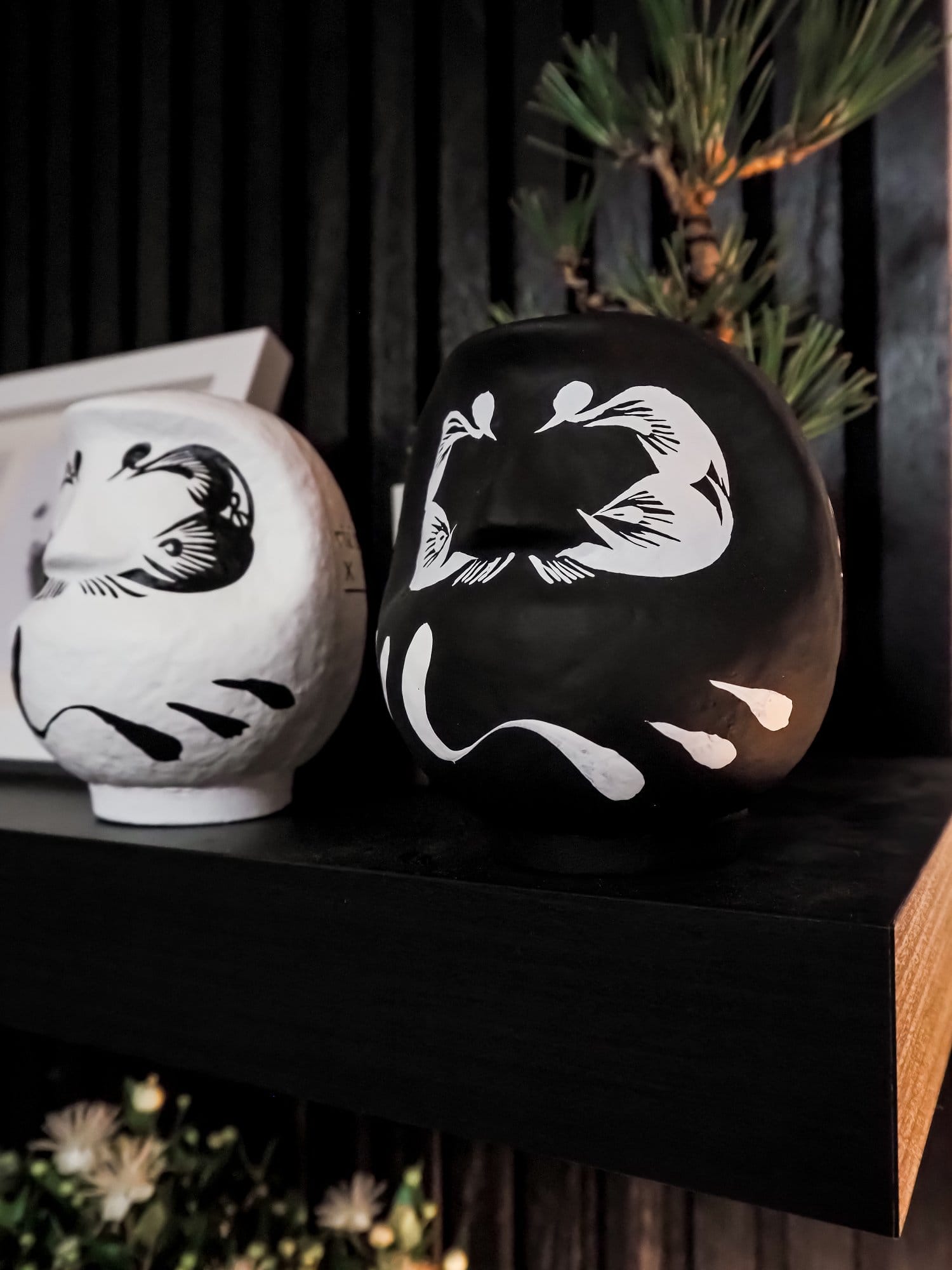 A close-up of two Daruma dolls, one black and one white, placed on a black shelf with a bonsai tree in the background