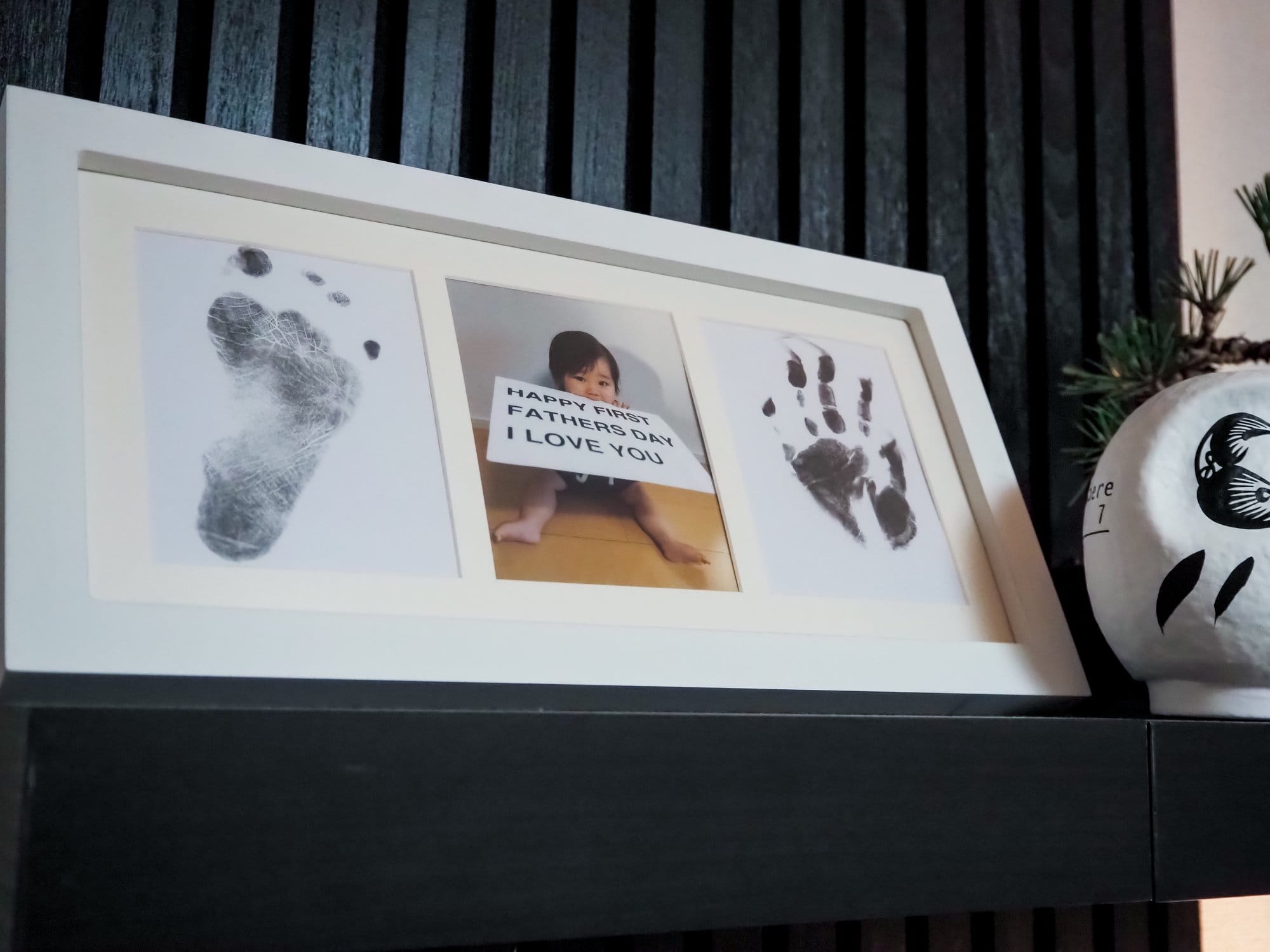 A close-up of a framed photo display featuring a baby’s footprint, a picture of the baby holding a “Happy First Father’s Day I Love You” sign, and a baby’s handprint, placed on a black shelf