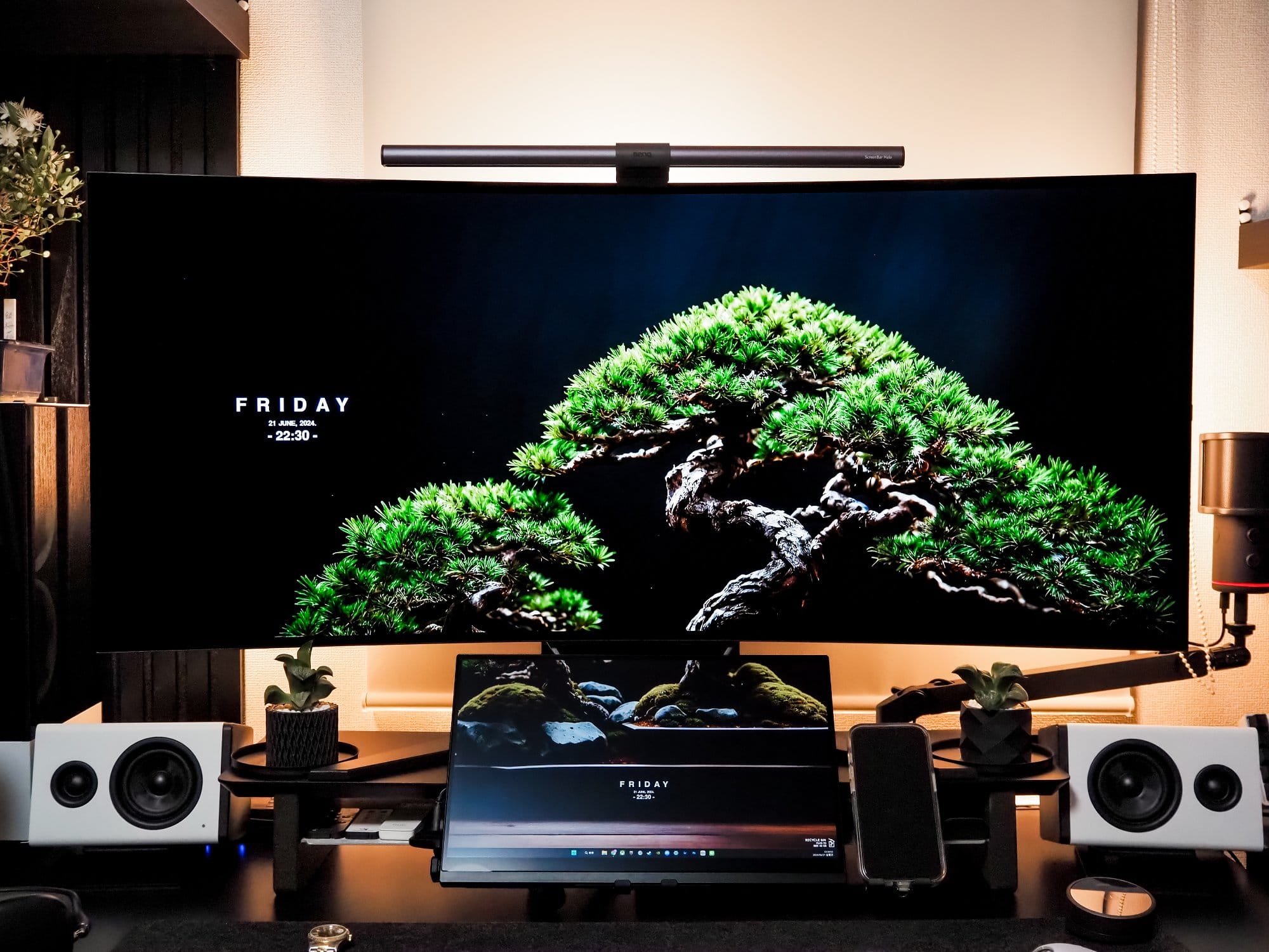 A well-organised gaming setup featuring a Corsair XENEON FLEX 45WQHD240 monitor with a BenQ ScreenBar Halo light bar, surrounded by bonsai trees, and white NZXT Relay speakers