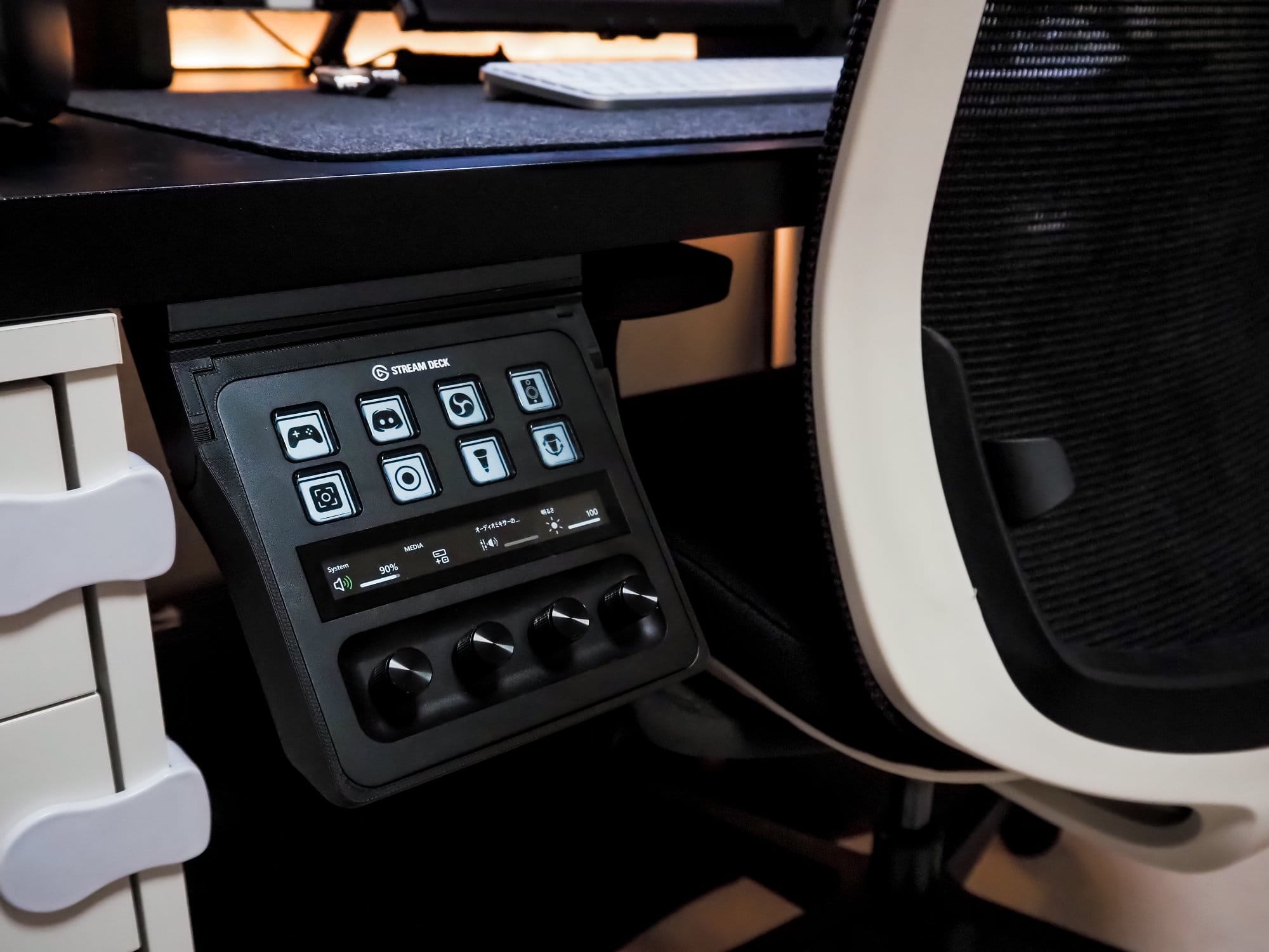 A close-up of an Elgato Stream Deck Plus mounted under a desk, next to a white gaming chair by AIMchair