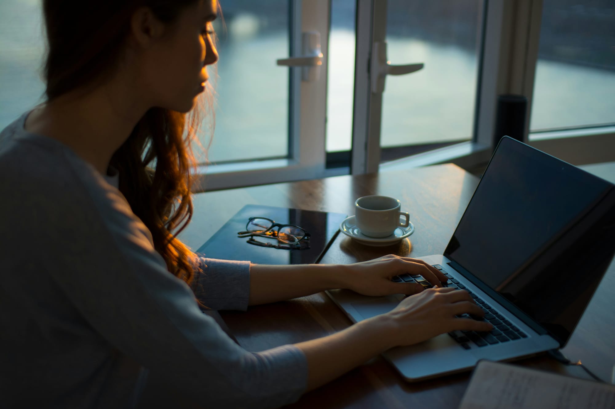 A woman working alone on a laptop at a wooden table by the open window