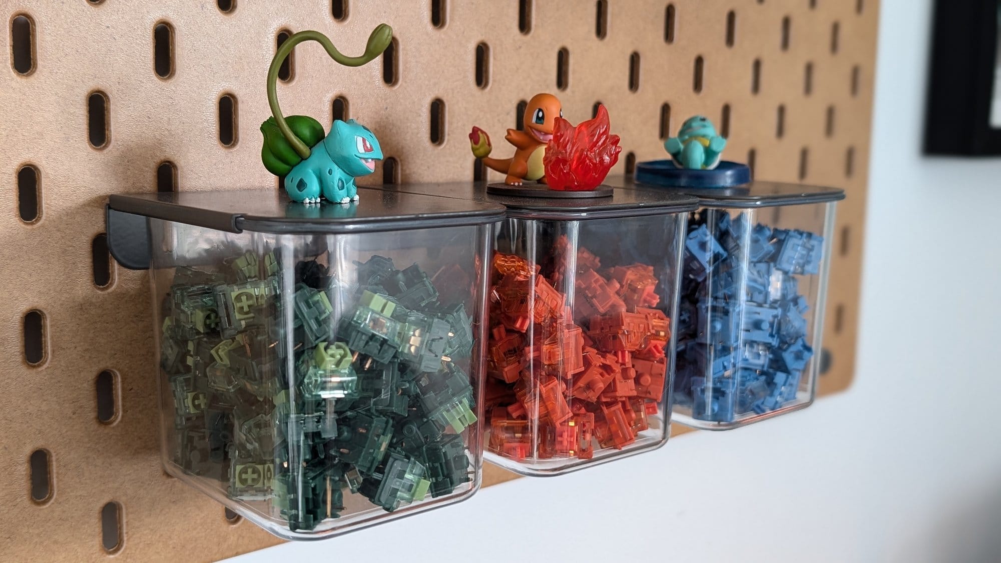 A well-organised pegboard setup displaying colourful mechanical keyboard switches in clear containers, each topped with a Pokémon figure