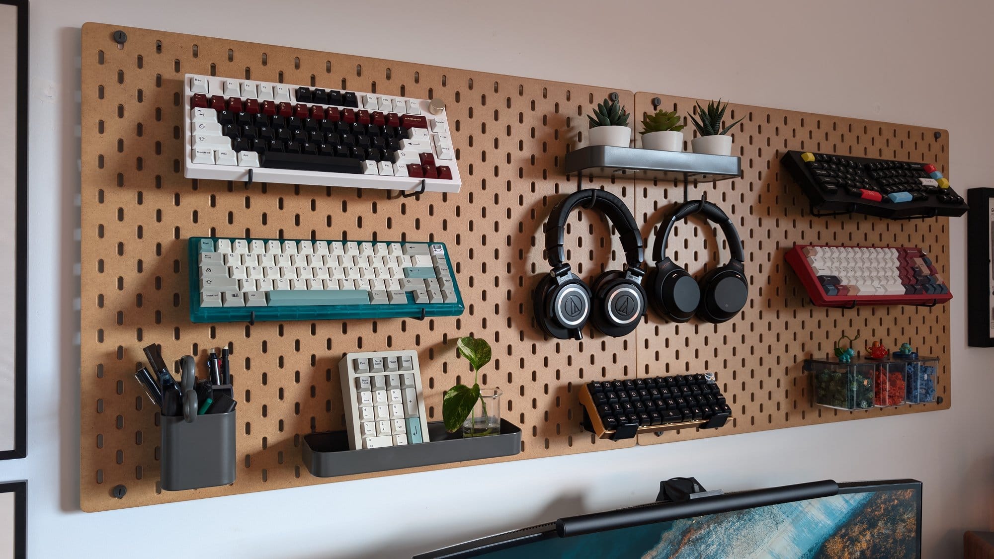 A well-organised pegboard display featuring a collection of mechanical keyboards, Audio Technica ATH-M50X and Sony WH-1000XM3 headphones, along with neatly arranged stationery and small plants