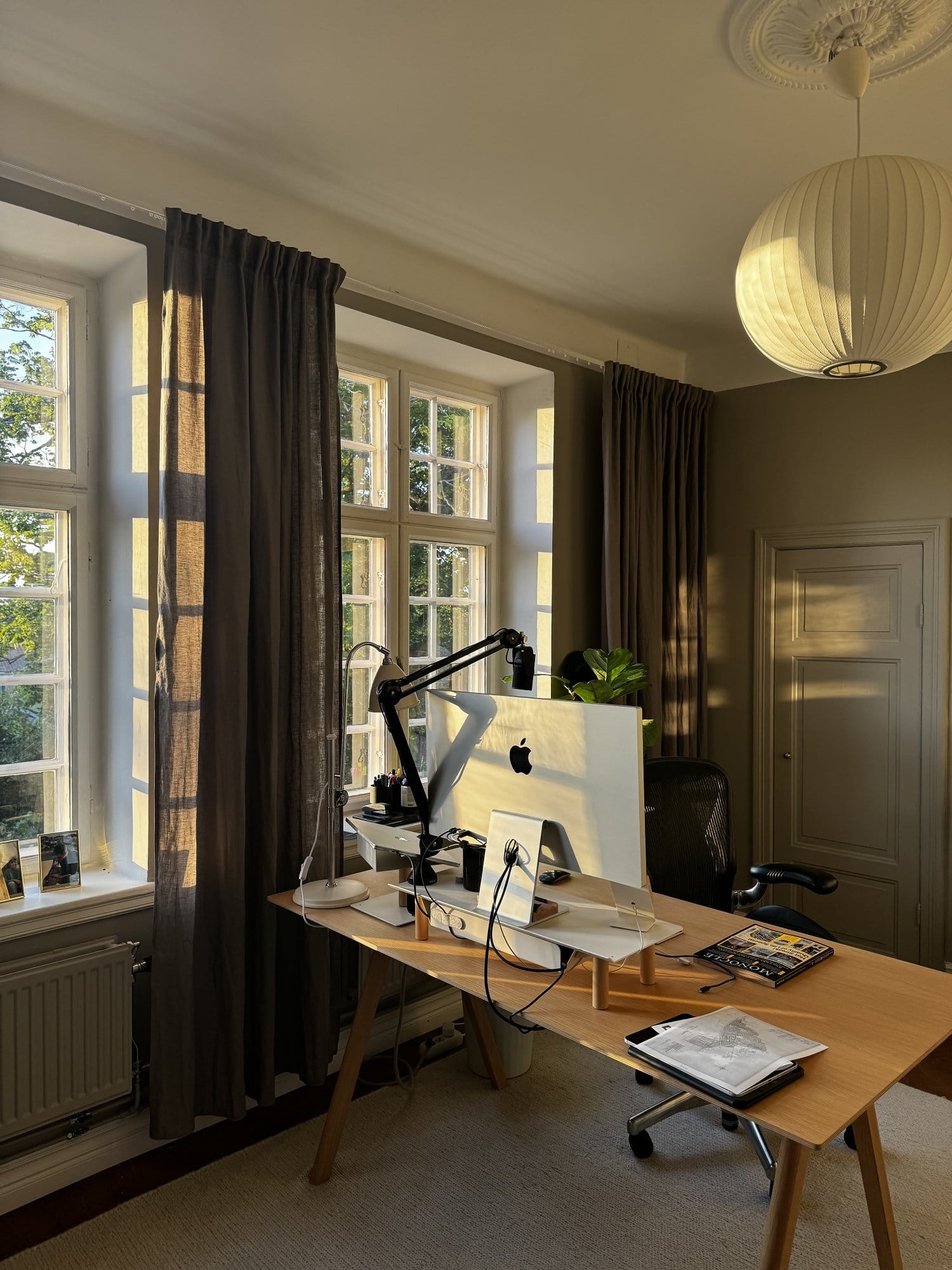 A warm and inviting home office featuring an Apple Studio Display on a wooden desk, bathed in natural light from large windows