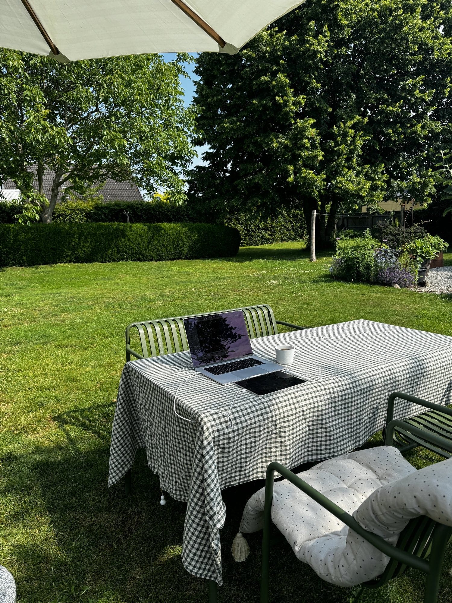 A serene outdoor workspace with a MacBook and iPad on a checkered tablecloth, set under a large umbrella in a lush green garden