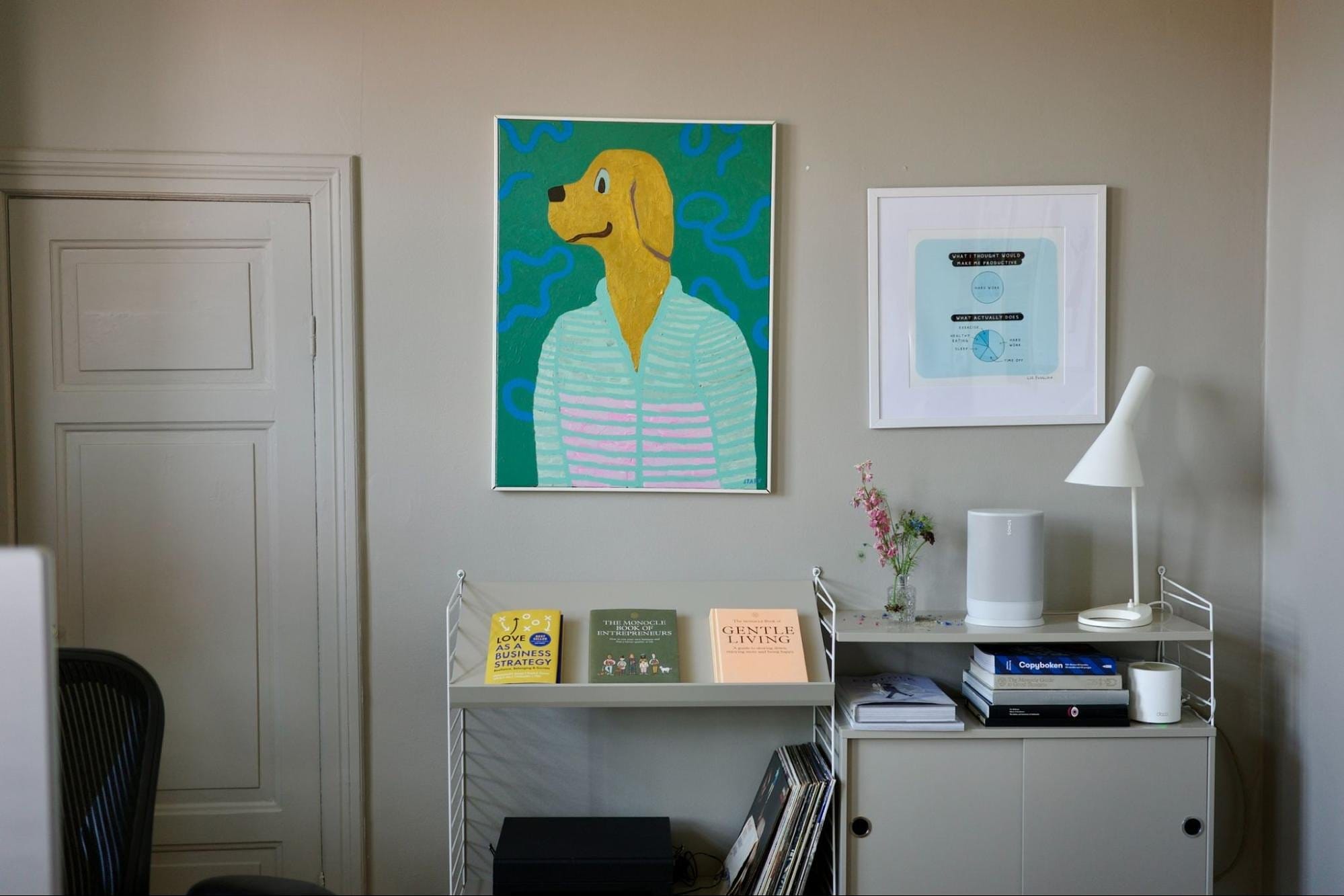 A thoughtfully arranged home office space featuring a vibrant yellow dog painting, a Bestlite BL1 desk lamp, and a Sonos Move speaker, with a shelf displaying a selection of books and a vase of flowers