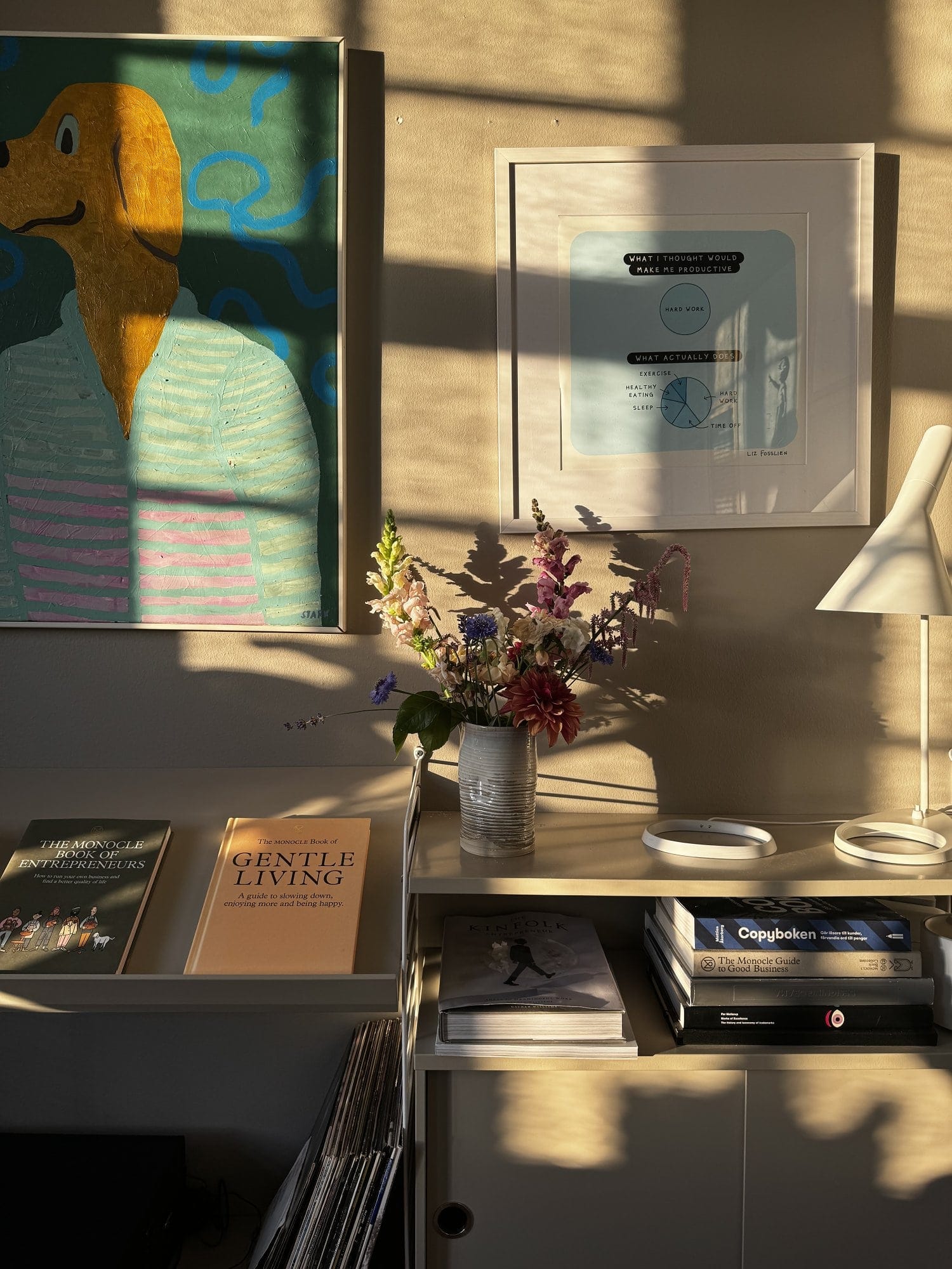 A cosy corner of the office featuring a Bestlite BL1 desk lamp, a vibrant dog painting, and sunlight casting warm shadows over a collection of books and a vase of flowers