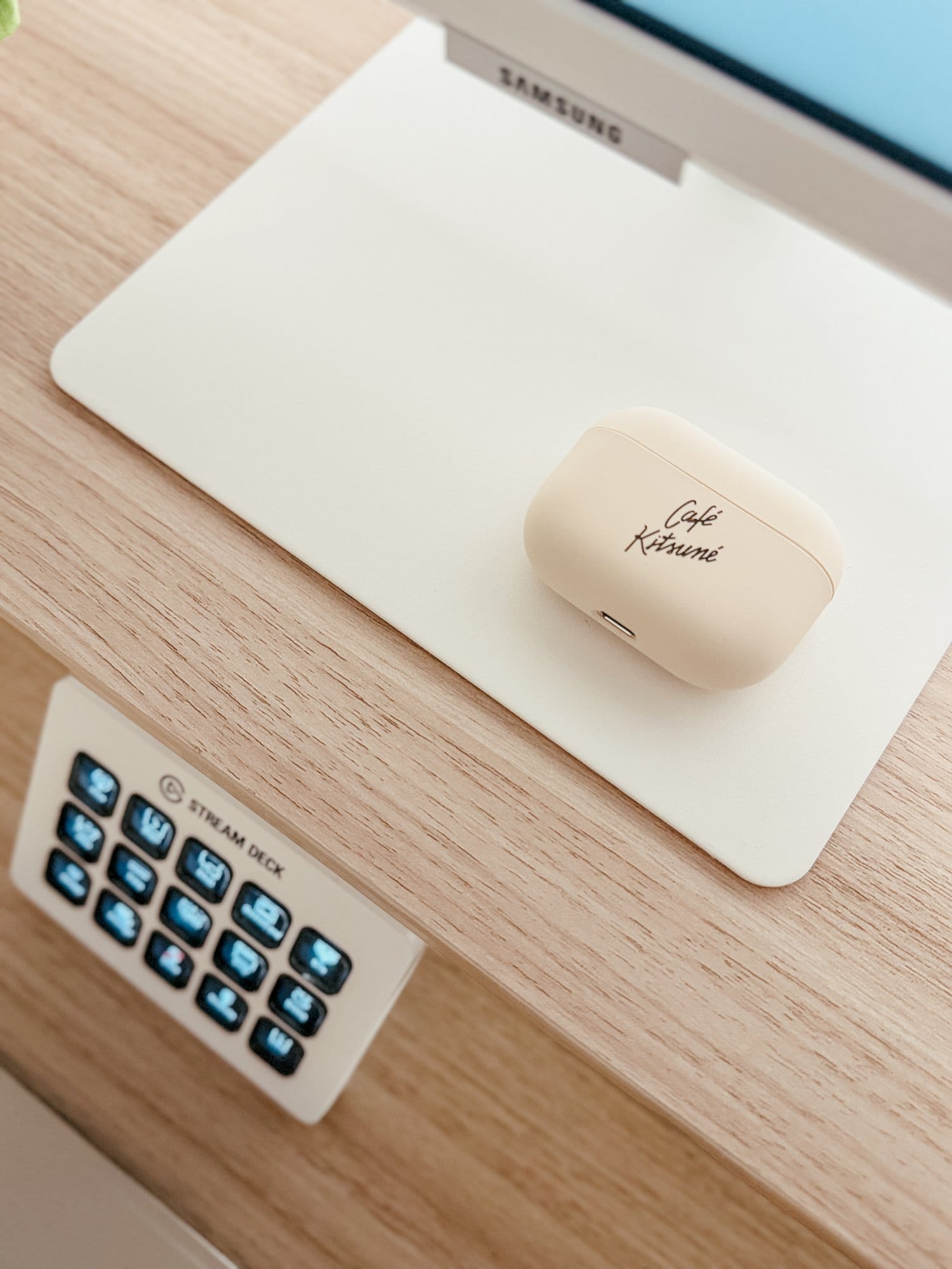 A close-up of the beige Café Kitsuné earbud case resting on a Samsung monitor stand, with a Stream Deck positioned below on a wooden desk