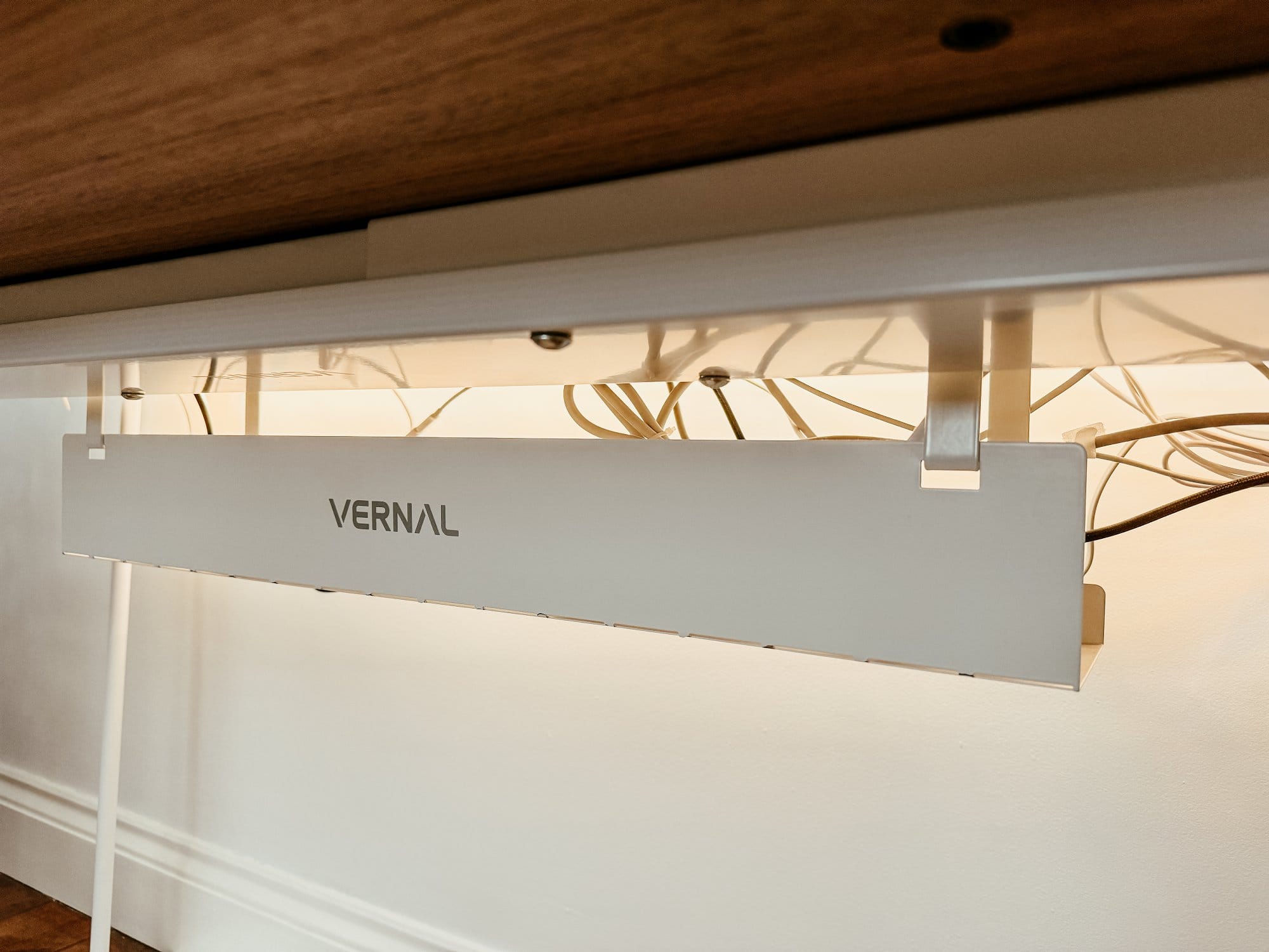 Under-desk cable management solution by Vernal, mounted beneath a wooden tabletop. The white cable tray is neatly organising and concealing numerous cables, providing a clean and clutter-free workspace