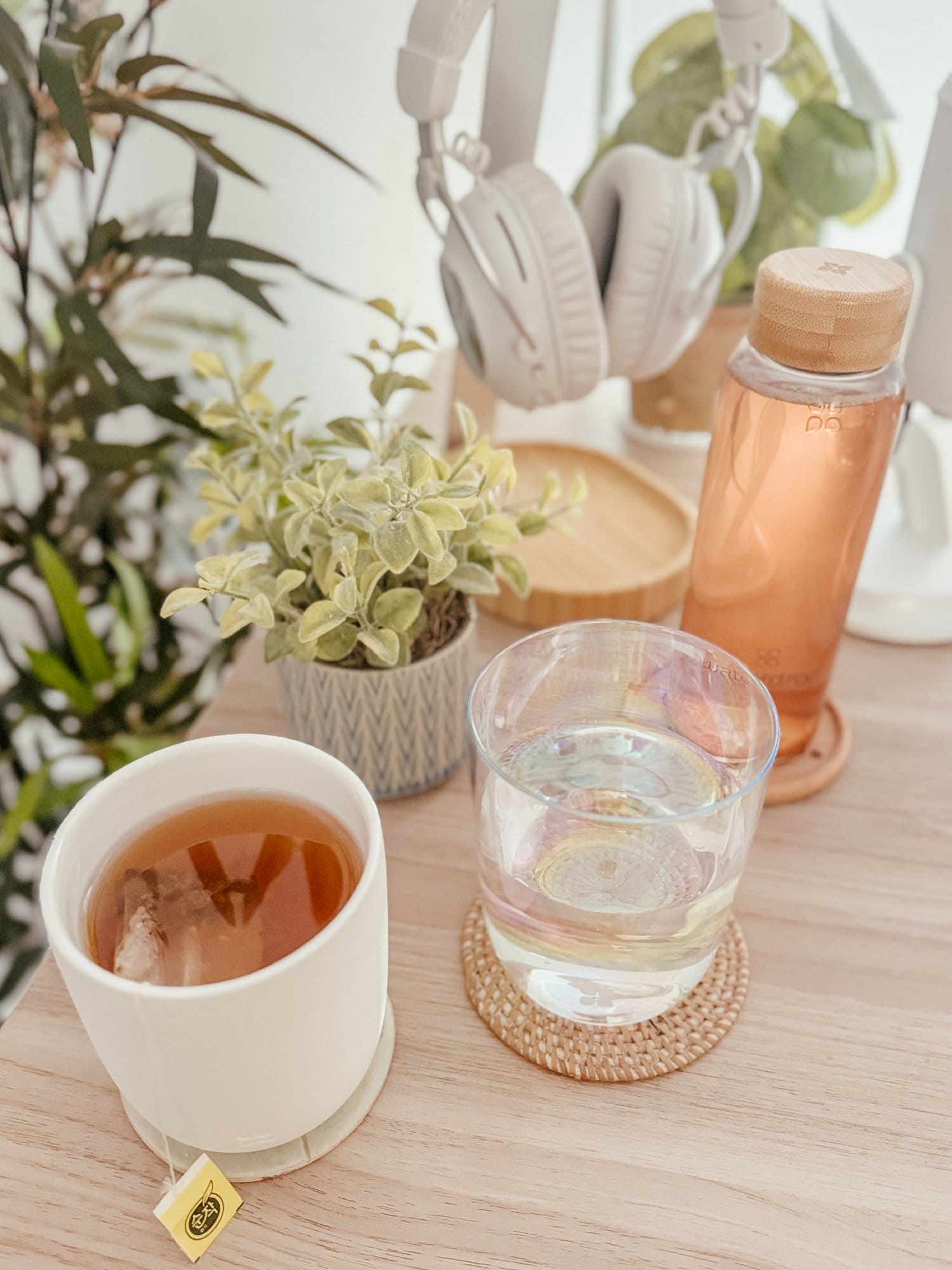 A refreshing desk setup featuring a cup of tea and a glass of water on woven coasters, accompanied by a potted plant, a bottle with a bamboo lid, and a pair of white headphones