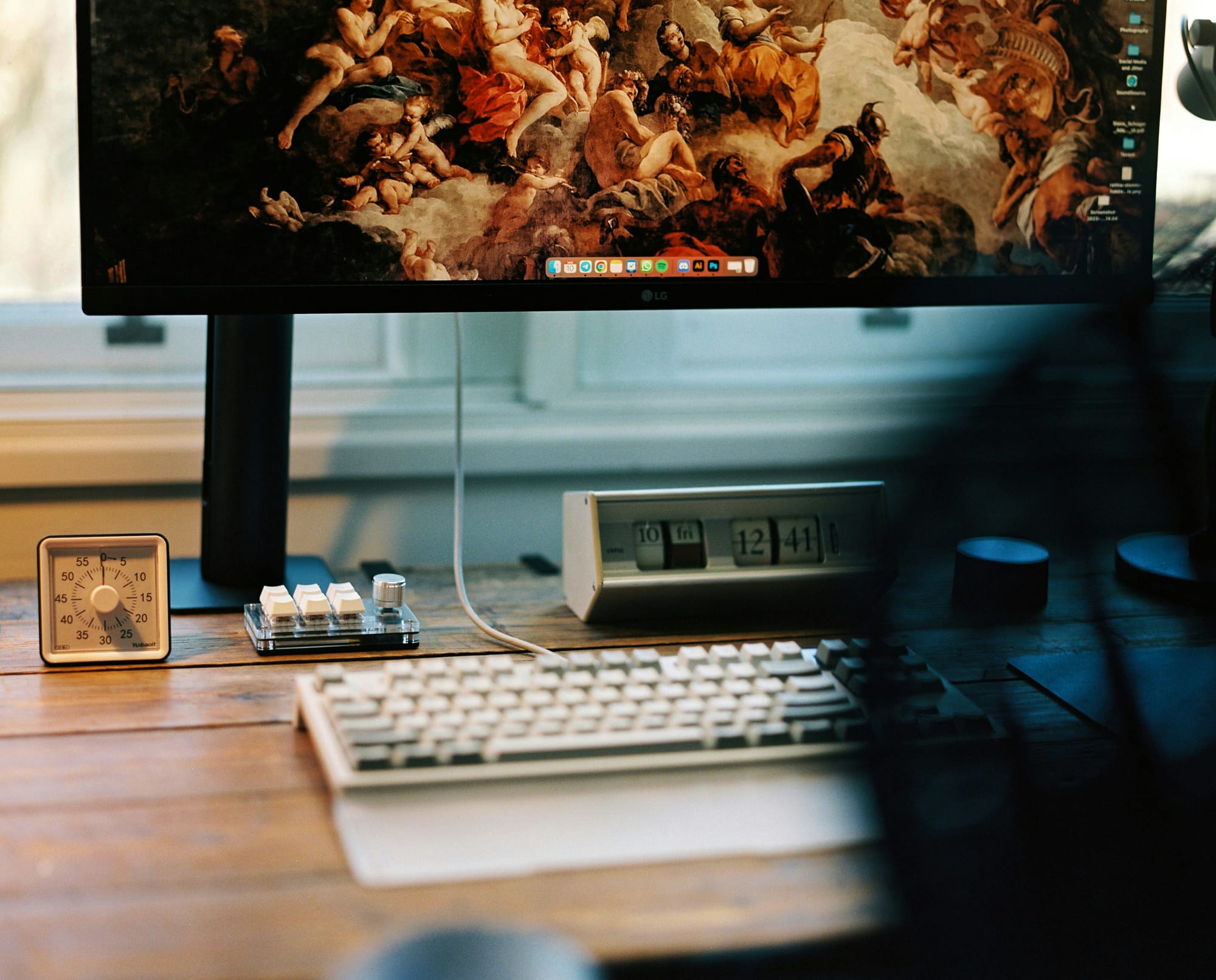 A window-facing desk setup with a monitor displaying a classical painting, a mechanical keyboard, and a retro flip clock