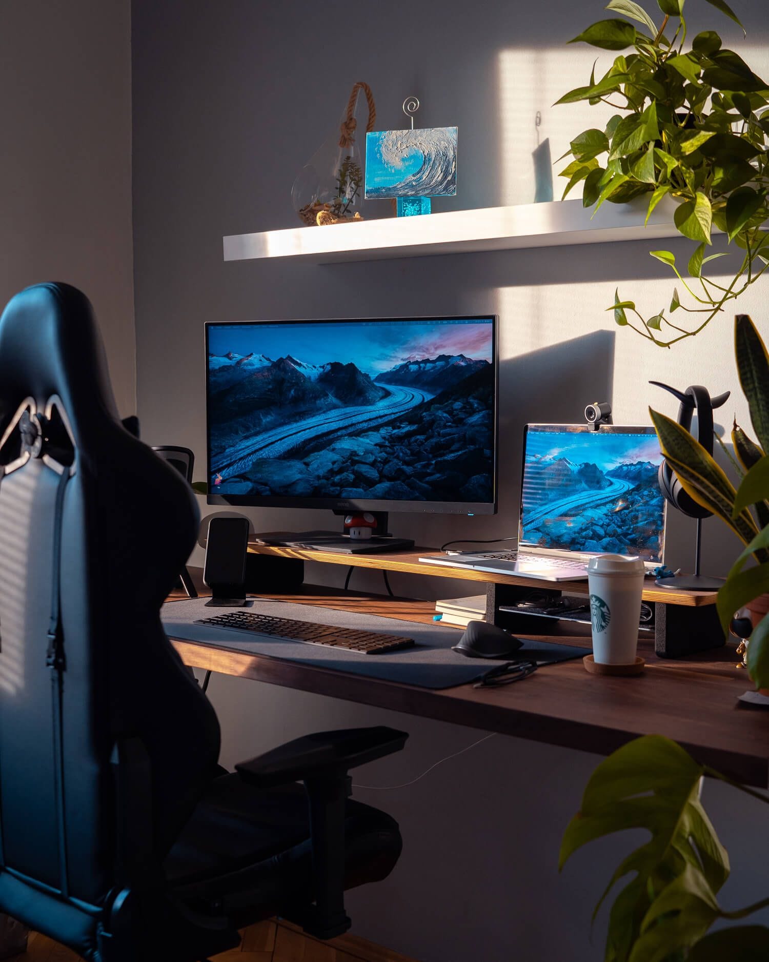 A stylish home desk setup featuring a gaming chair, a monitor, a laptop, and a wood-toned desk adorned with plants and a Starbucks cup