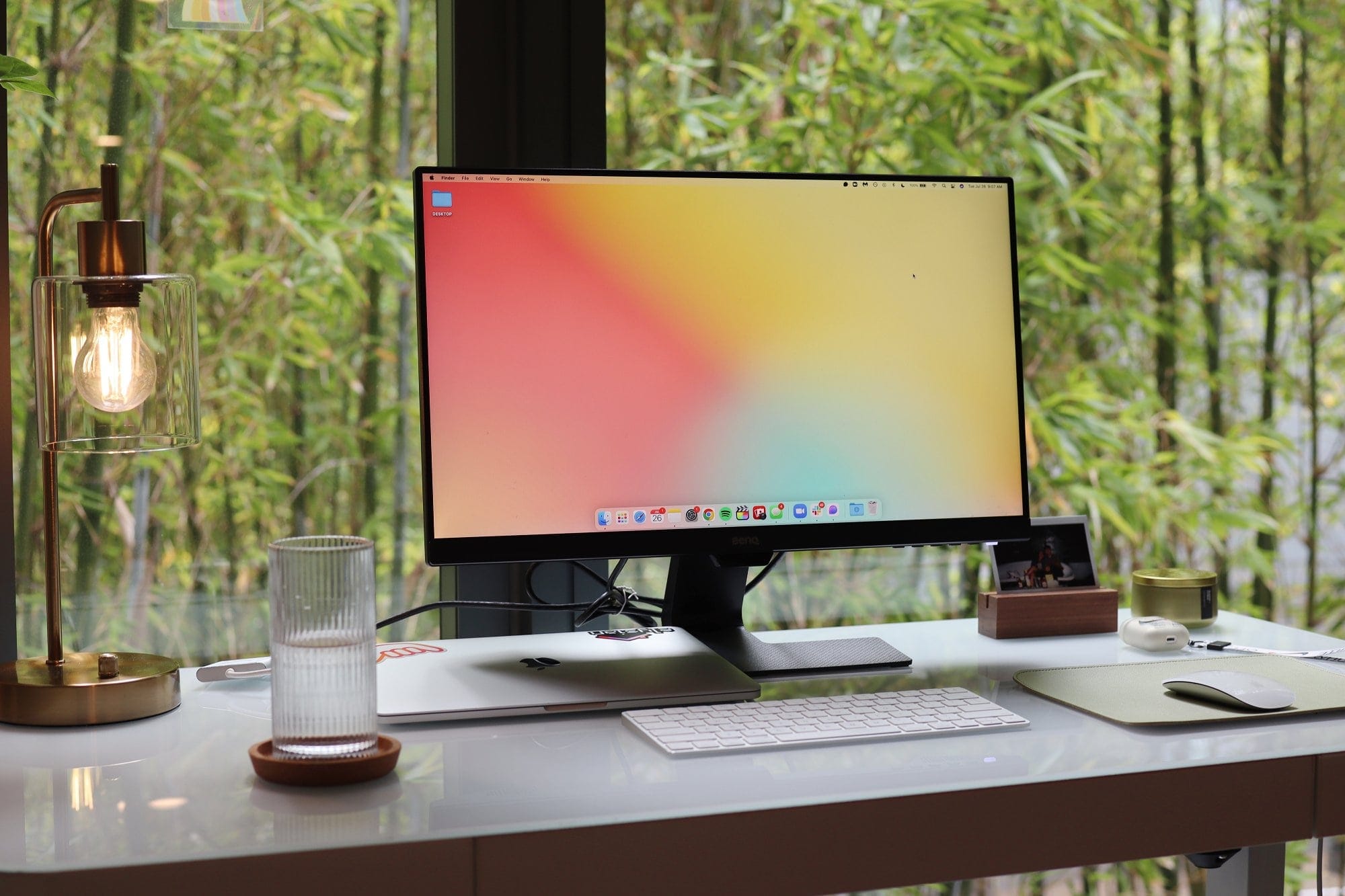 A serene and modern workspace featuring a large monitor with a minimalist desktop setup, including a sleek keyboard, mouse, and a glass of water, all set against a backdrop of lush green bamboo visible through a large window