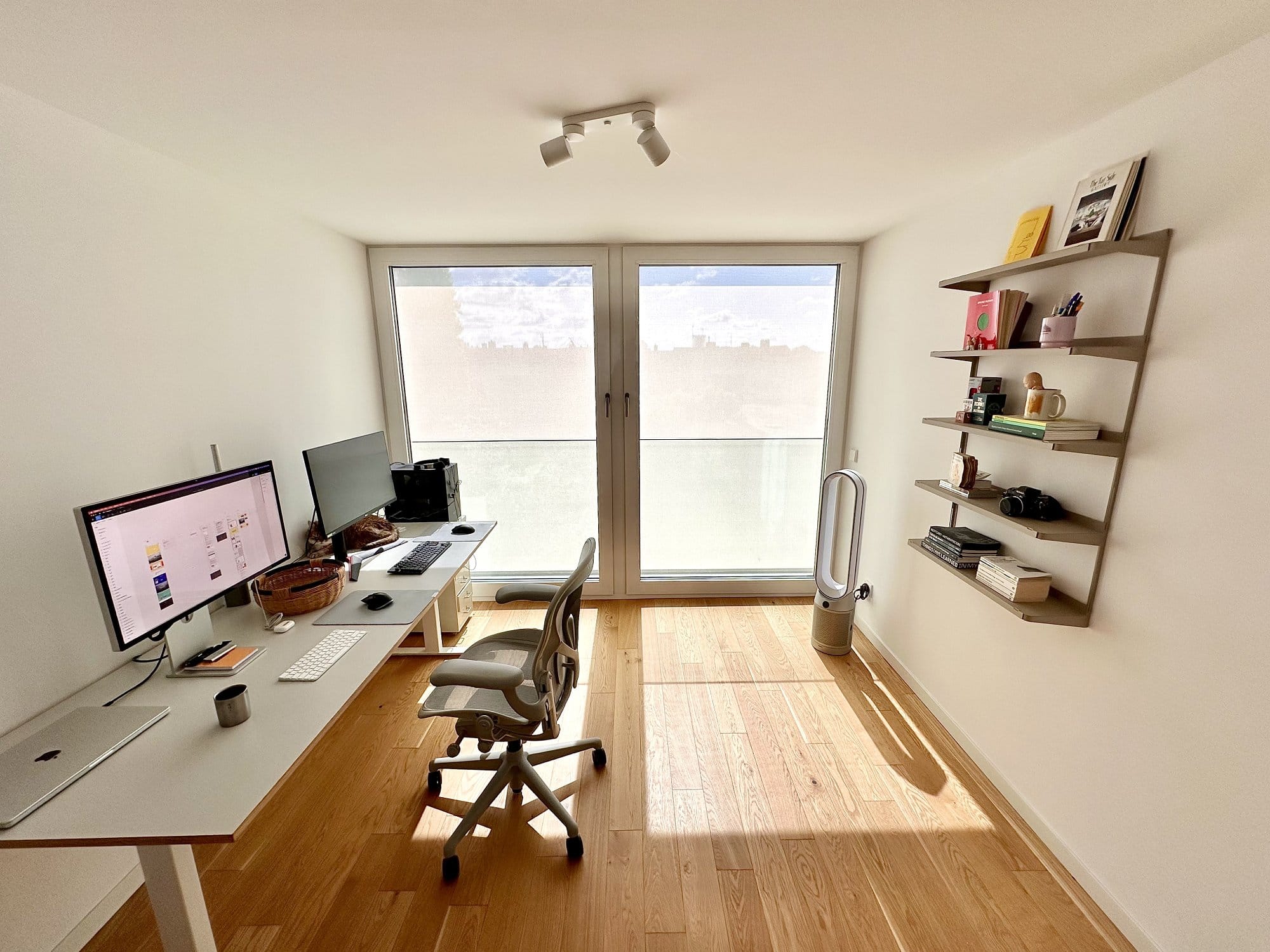 A bright and minimalist workspace featuring an Apple Studio Display and a MacBook Pro M2 Max on an automatic standing desk, complemented by a Herman Miller Aeron chair and a Dyson TP07 Purifier Cool