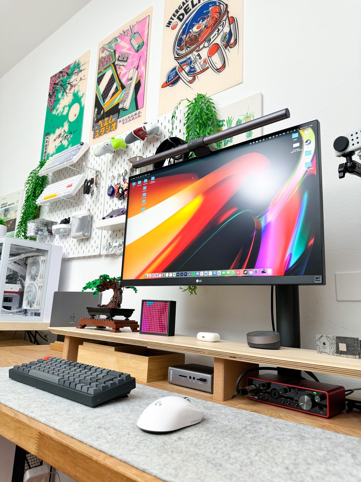 A modern workspace featuring an LG 27UN880P monitor with a BenQ monitor light bar, NuPhy Halo keyboard, Logitech G Superlight Pro mouse on a Harber London wool-like desk mat, MacBook Pro M1 Pro 2022, Focusrite audio card, and decorative bonsai, all arranged on an IKEA KARLBY tabletop with Flexispot legs, complemented by colourful wall art and various tech accessories on a pegboard