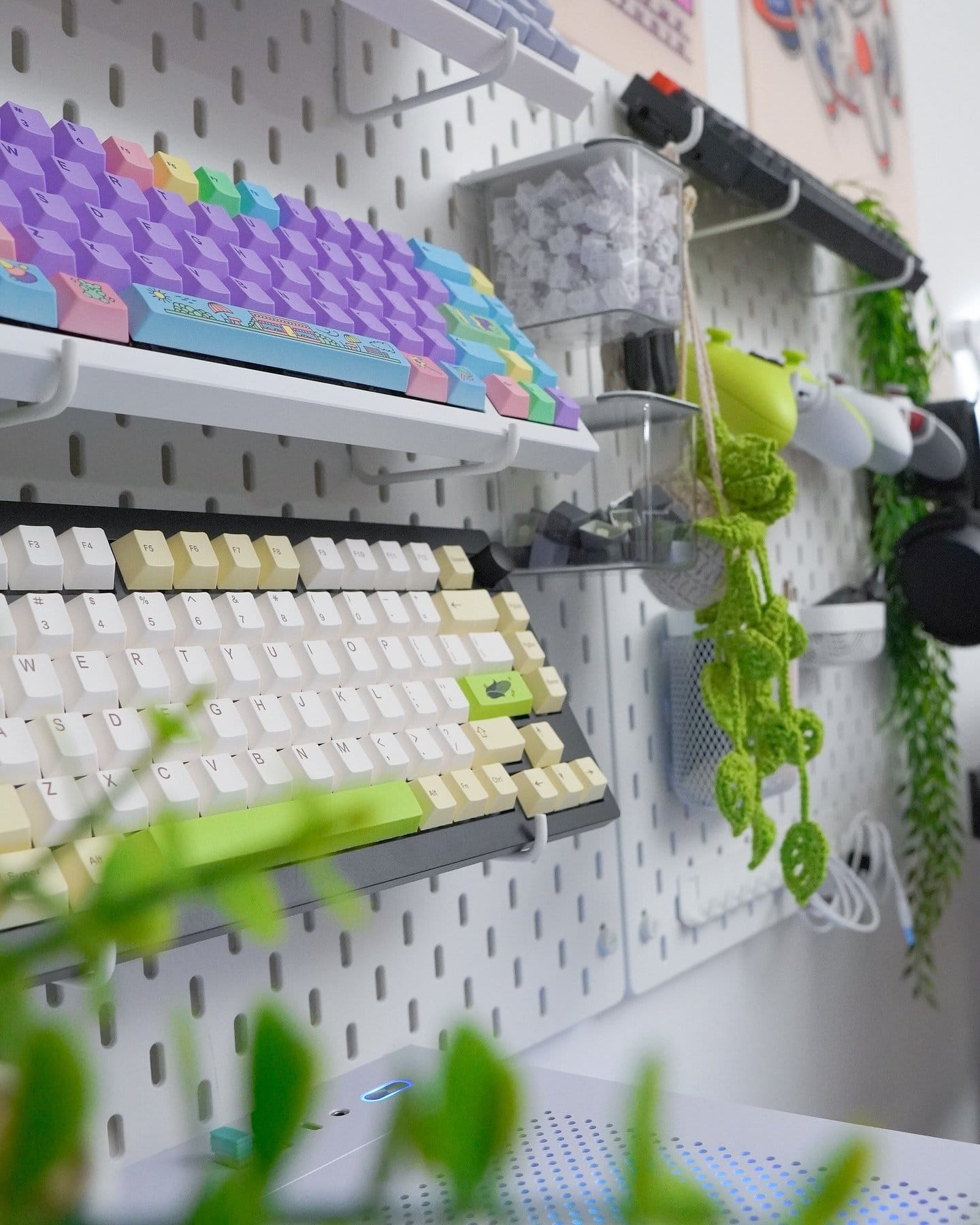 A close-up view of a wall-mounted pegboard displaying colourful NuPhy Halo mechanical keyboards, keycaps, and various tech accessories, with decorative green plants interspersed among the items