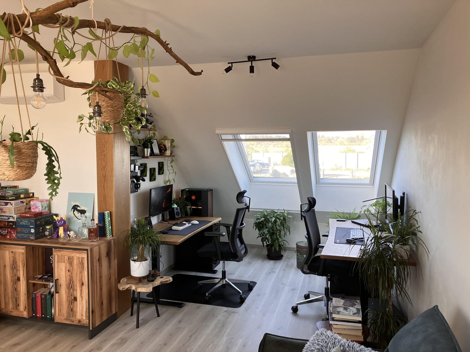 A spacious and creatively designed home office featuring two ergonomic workstations with large monitors, surrounded by lush indoor plants and natural light from skylights, with a ceiling-mounted wooden branch holding hanging plants and light fixtures