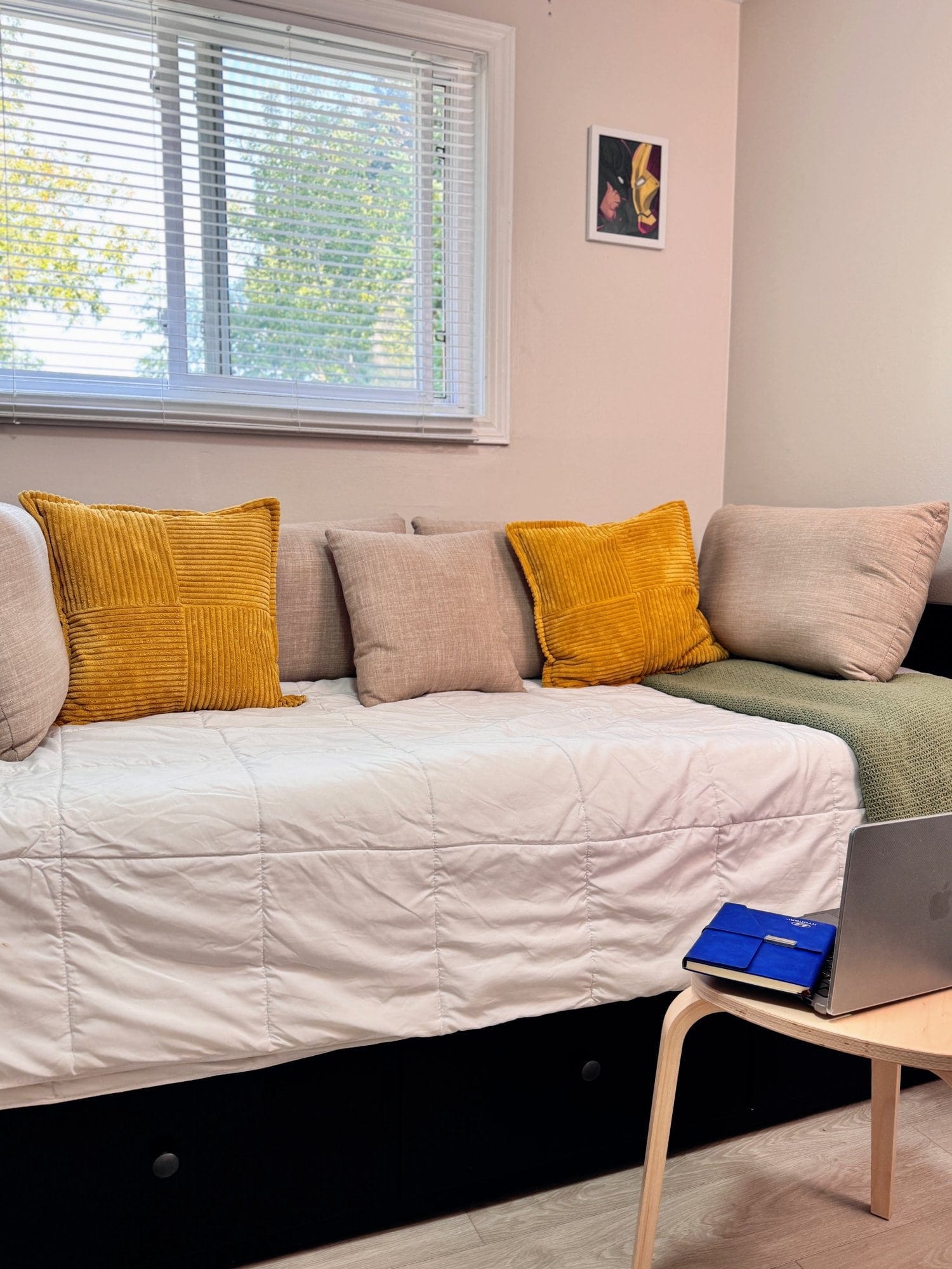  A cosy daybed setup with mustard yellow and beige cushions, a soft white duvet, and a small side table holding a laptop and notebook