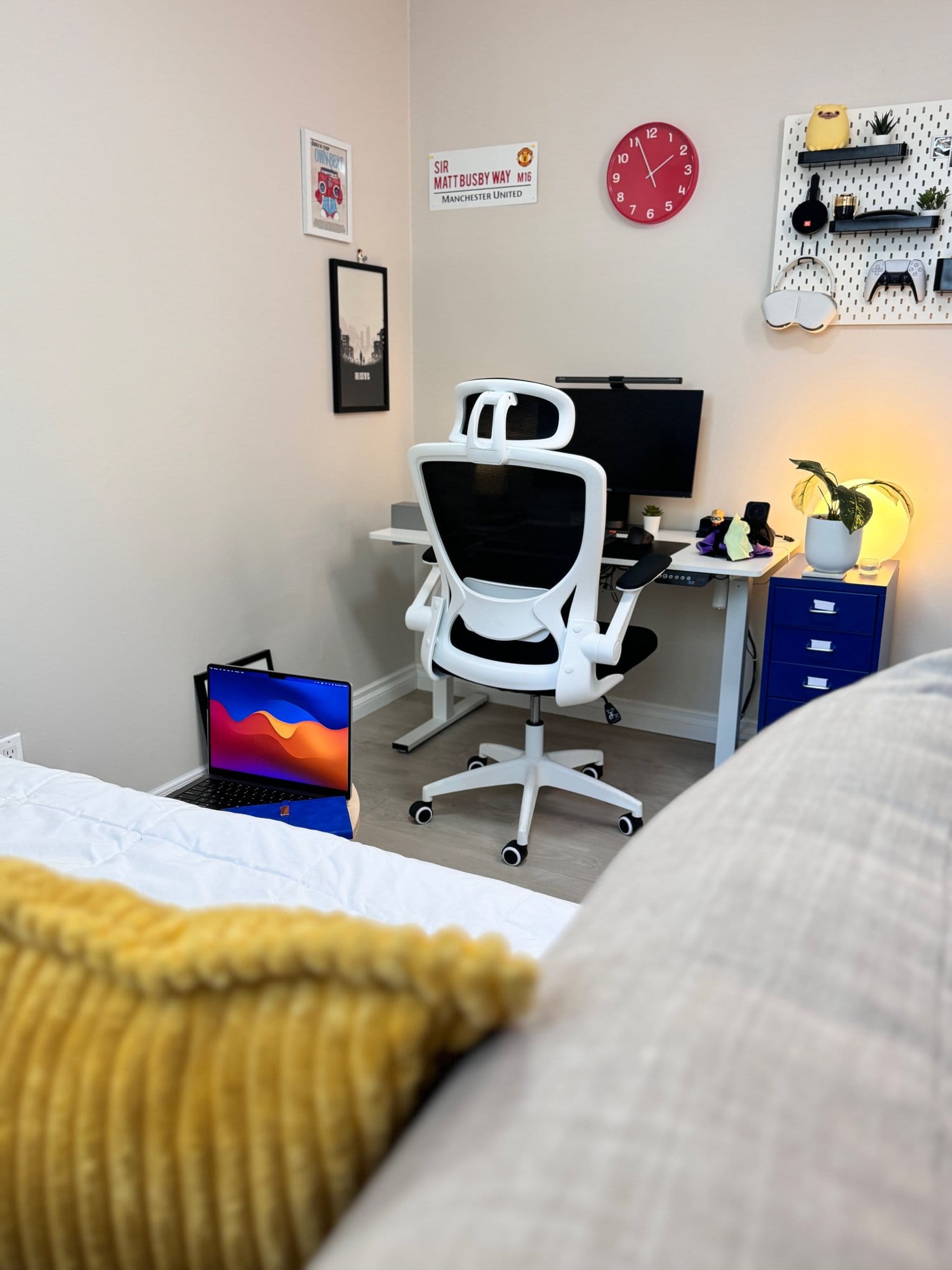 A home office corner with a white ergonomic KERDOM chair at a minimalist desk setup and a MacBook Pro 2023 on the floor near a cosy daybed adorned with mustard yellow cushions