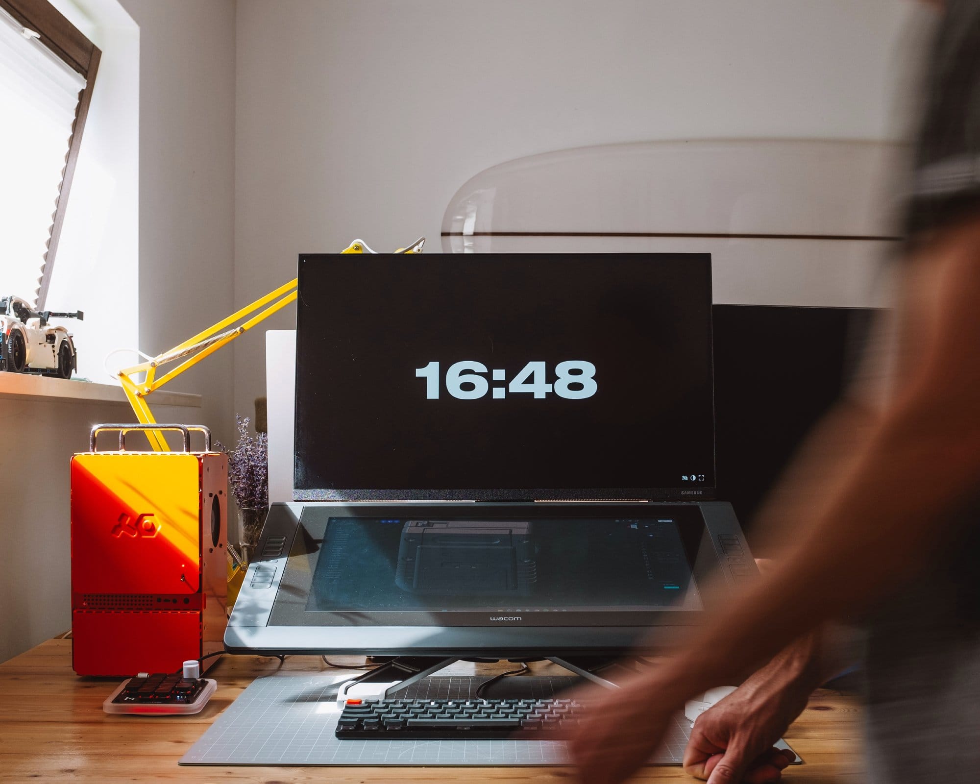 A modern desk setup with a Samsung Space monitor displaying the time, a Wacom Cintiq 22HD tablet below it, a yellow lamp, and an orange Computer-1 (TE) case on the left