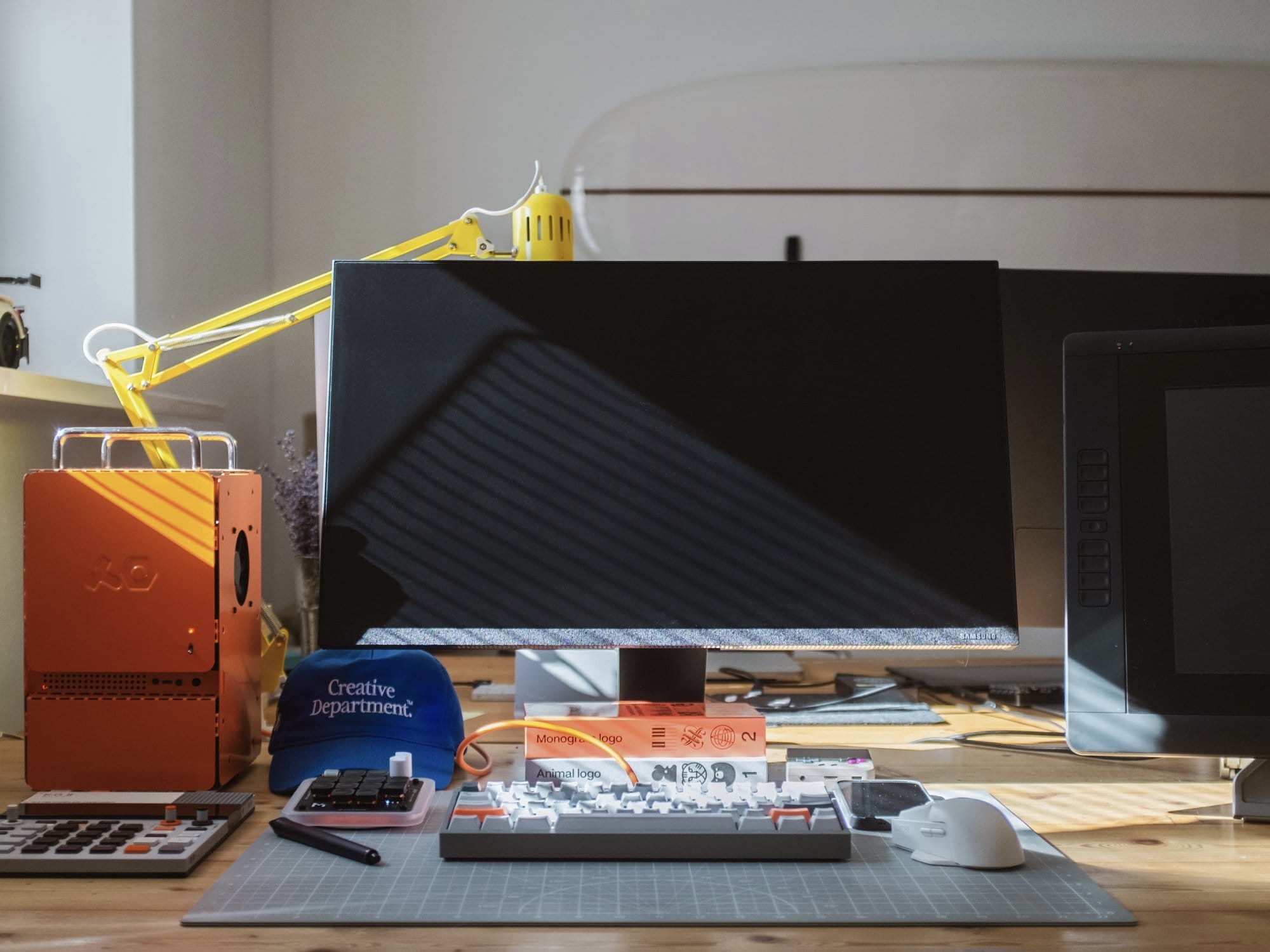  A tidy workspace with a central Samsung Space monitor, flanked by a Wacom Cintiq 22HD tablet, an orange Computer-1 (TE) case, and a yellow lamp, with a custom-built keyboard and a “Creative Department” cap