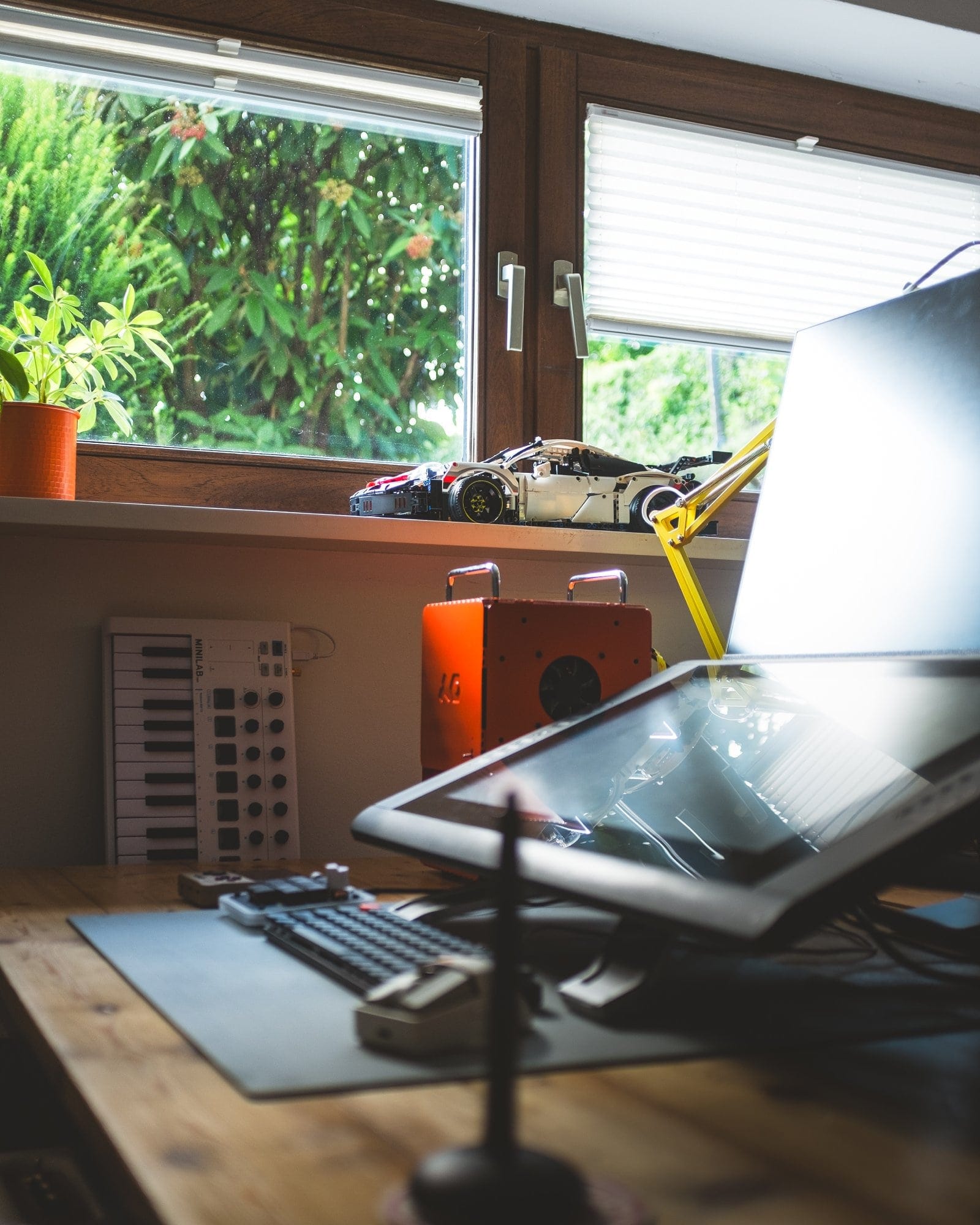 A cosy desk setup with a Wacom Cintiq 22HD tablet, a custom-built keyboard, and a vibrant orange Computer-1 (TE) case, with a LEGO car and a MIDI keyboard placed on the windowsill next to a leafy green plant