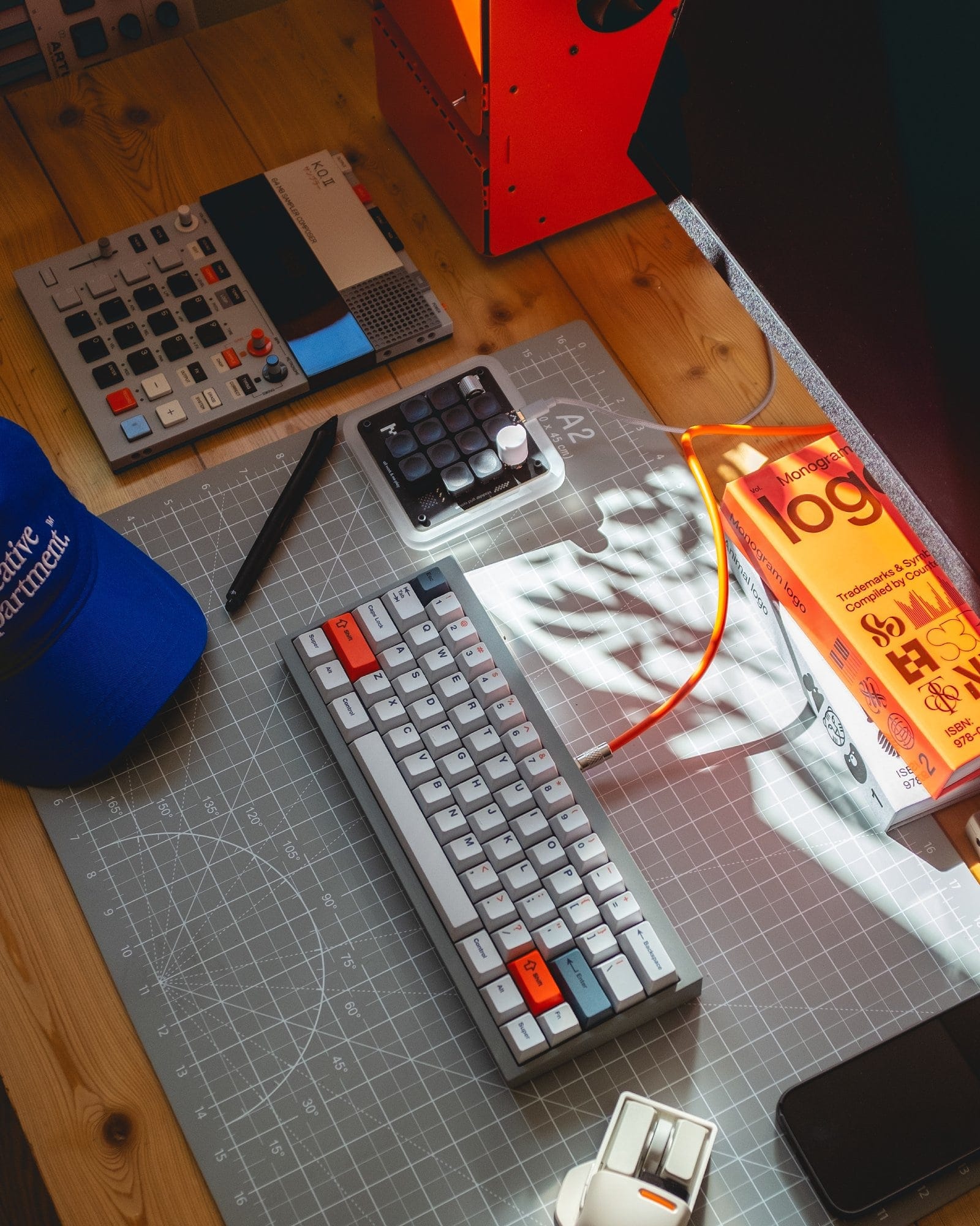 A well-organised workspace featuring a custom-built keyboard with orange and grey keys, a Work Louder macropad, design books, and creative tools like a KO2 instrument and pen