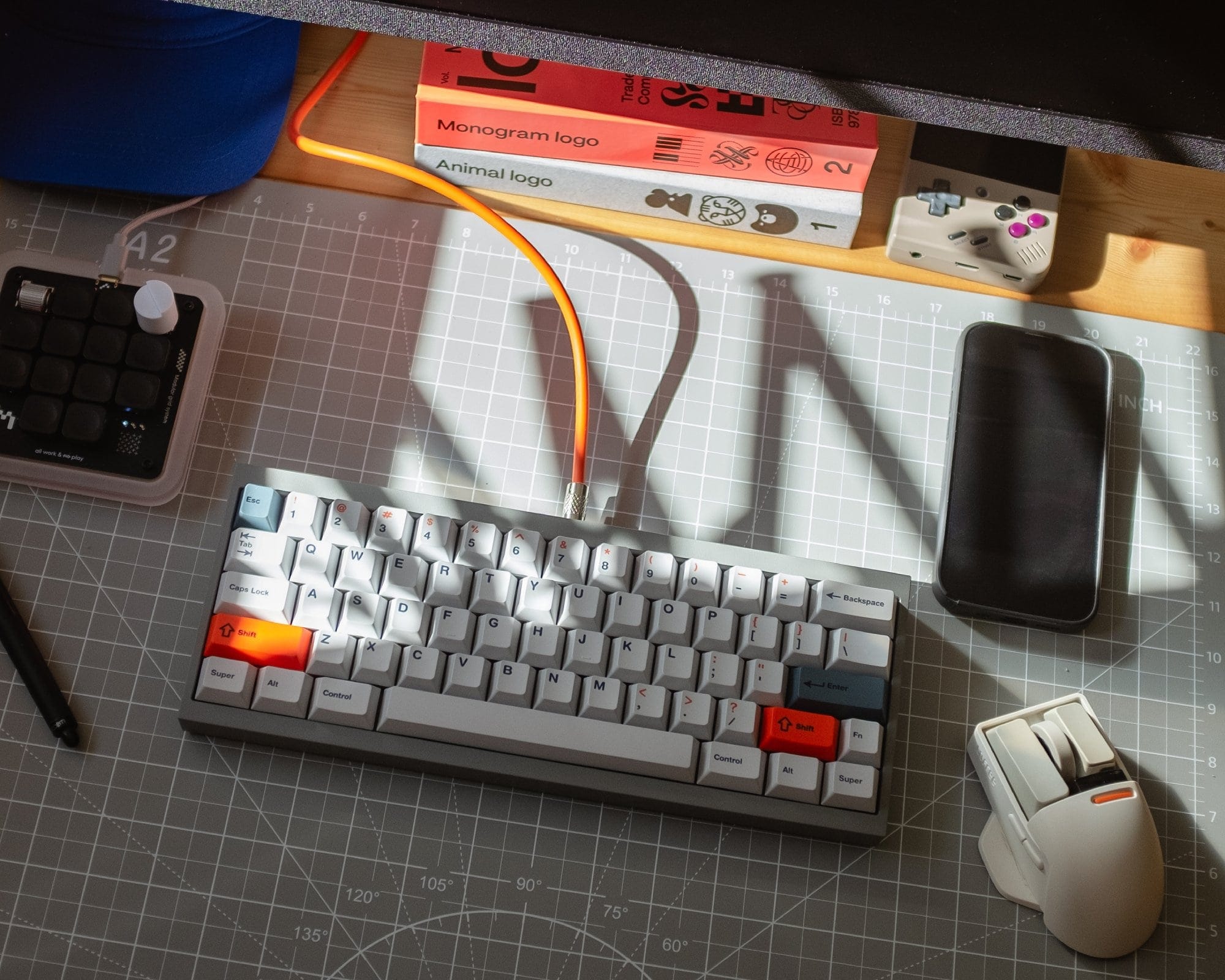 An organised desk setup featuring a custom-built keyboard with orange accents, a Work Louder macropad, a gaming mouse, and design books in the background