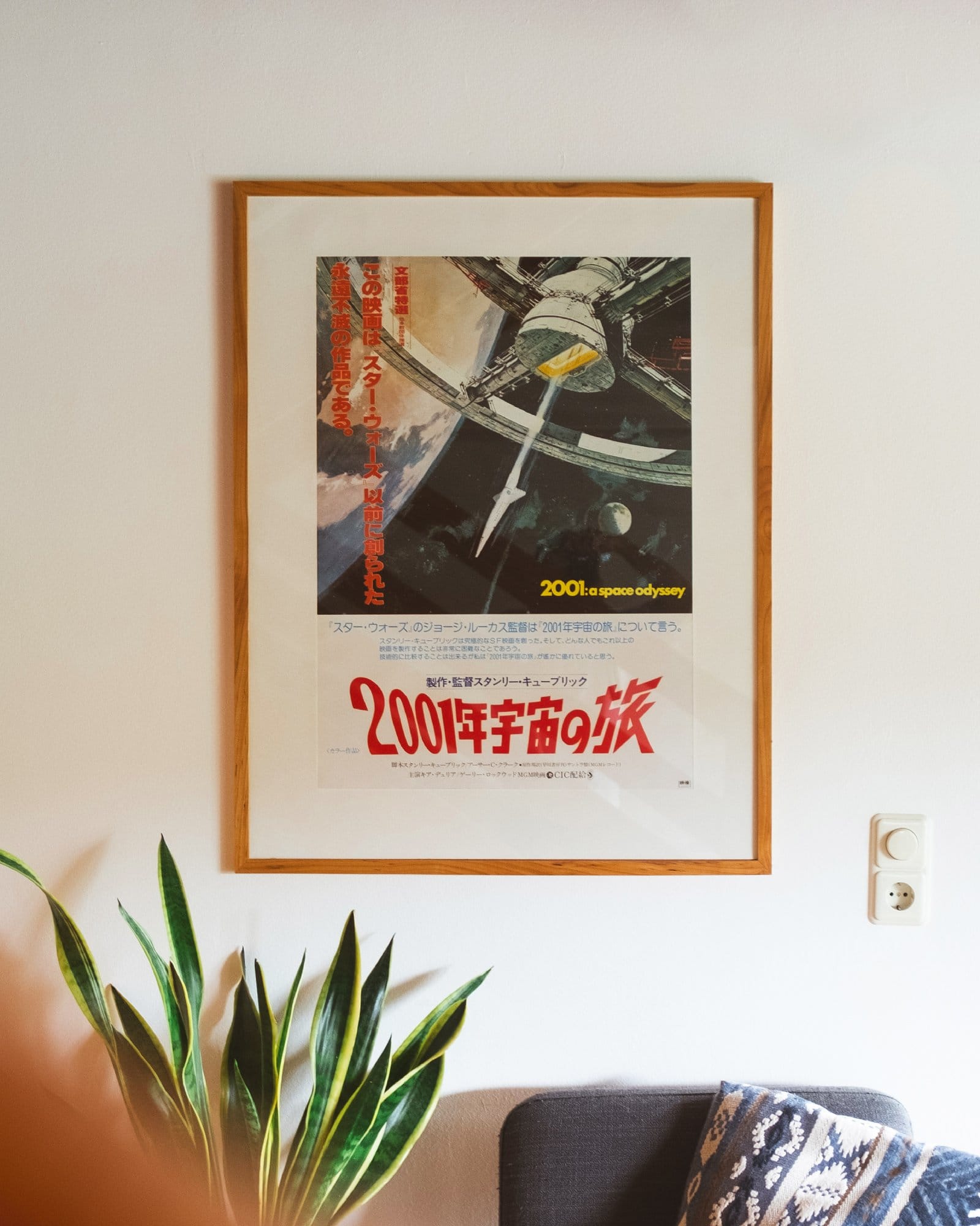 A framed Japanese poster of 2001: A Space Odyssey hangs on a white wall above a cosy corner with a patterned cushion and a green potted plant