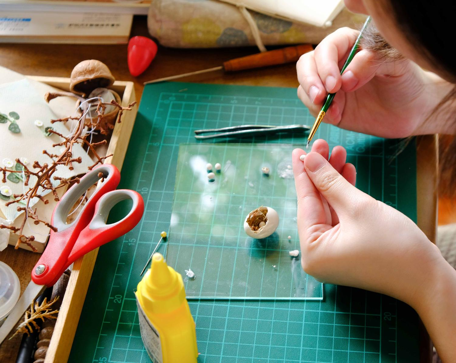 A close-up of a person carefully working on a delicate craft project, using a small brush and tweezers, with scattered tools such as scissors, glue, and natural materials on a cutting mat