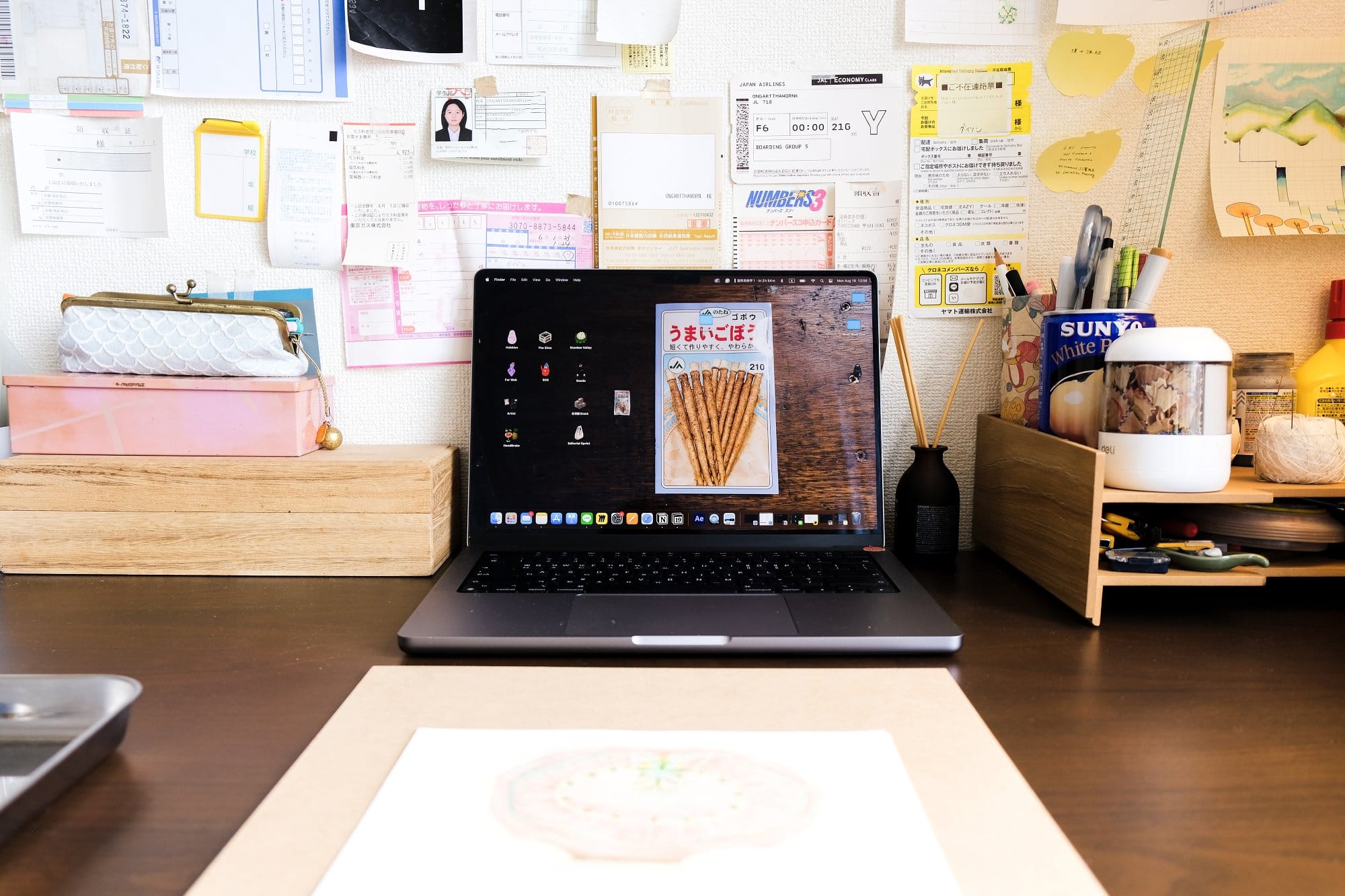 A laptop-only desk setup with a MacBook displaying a snack image as the desktop wallpaper, surrounded by organised stationery, a pink box, a wooden organiser, and a wall filled with notes and papers