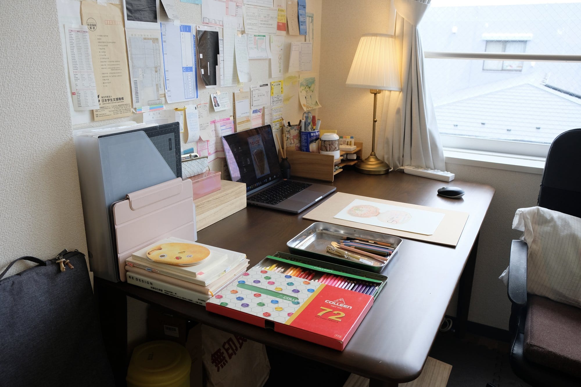 A neatly arranged desk with art supplies, a laptop, and sketchbooks, alongside a tray of pencils, set near a window with natural light and an IKEA ÅRSTID lamp providing additional lighting