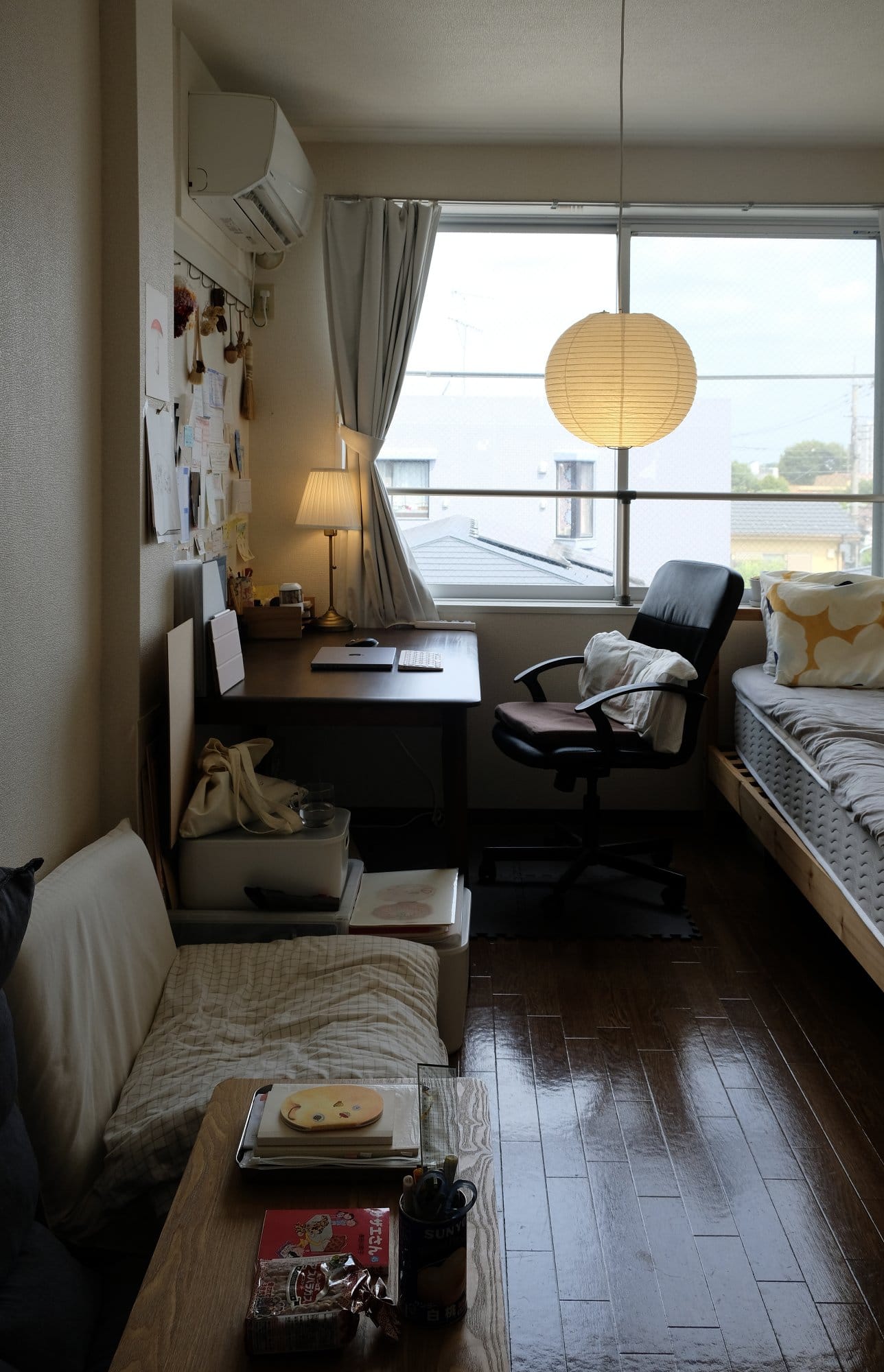 A small, organised room with a desk by the window, featuring a chair, lamp, and workspace, with a sofa and bed positioned close together in the compact space