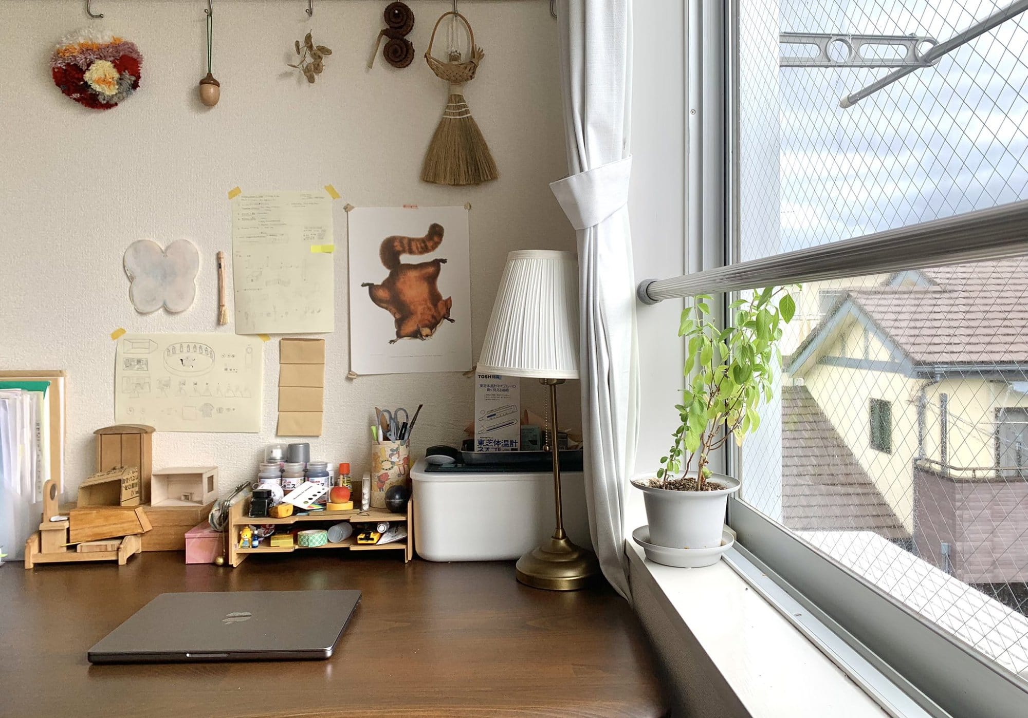 A desk with a closed laptop, a wooden organiser holding supplies, a lamp by the window, and a small plant on the windowsill, with wall decorations and drawings above