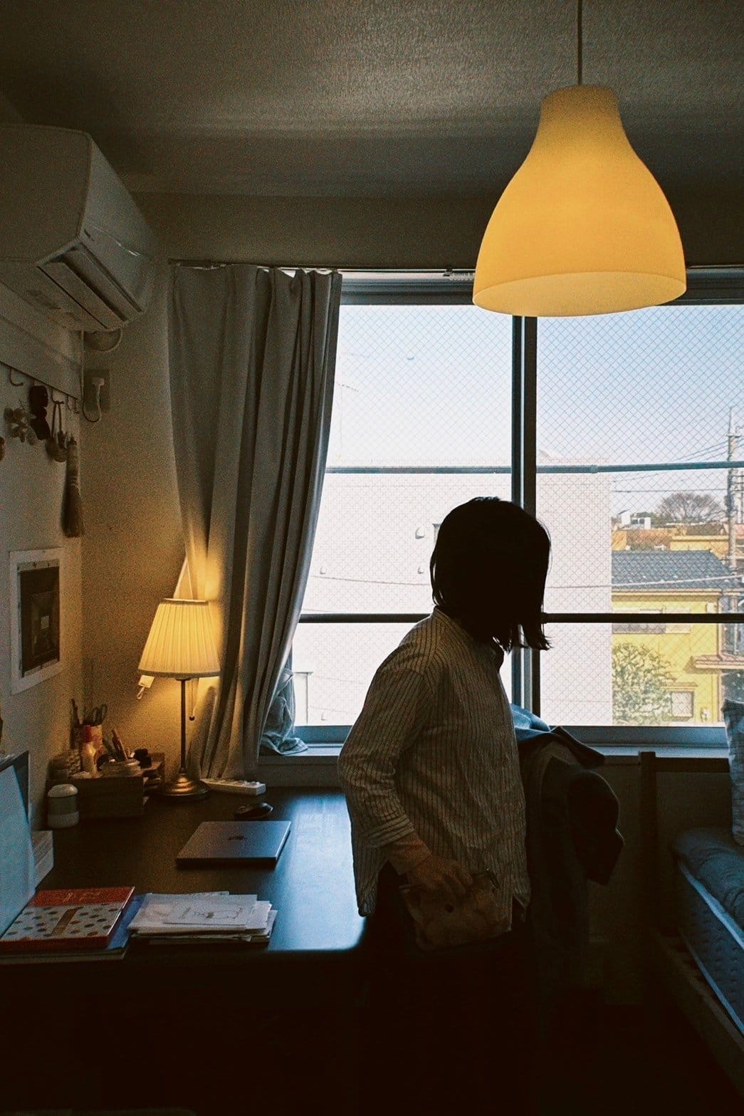 A person stands in a softly lit room, near a desk with a lamp and books, with natural light coming through the window and a ceiling light above them