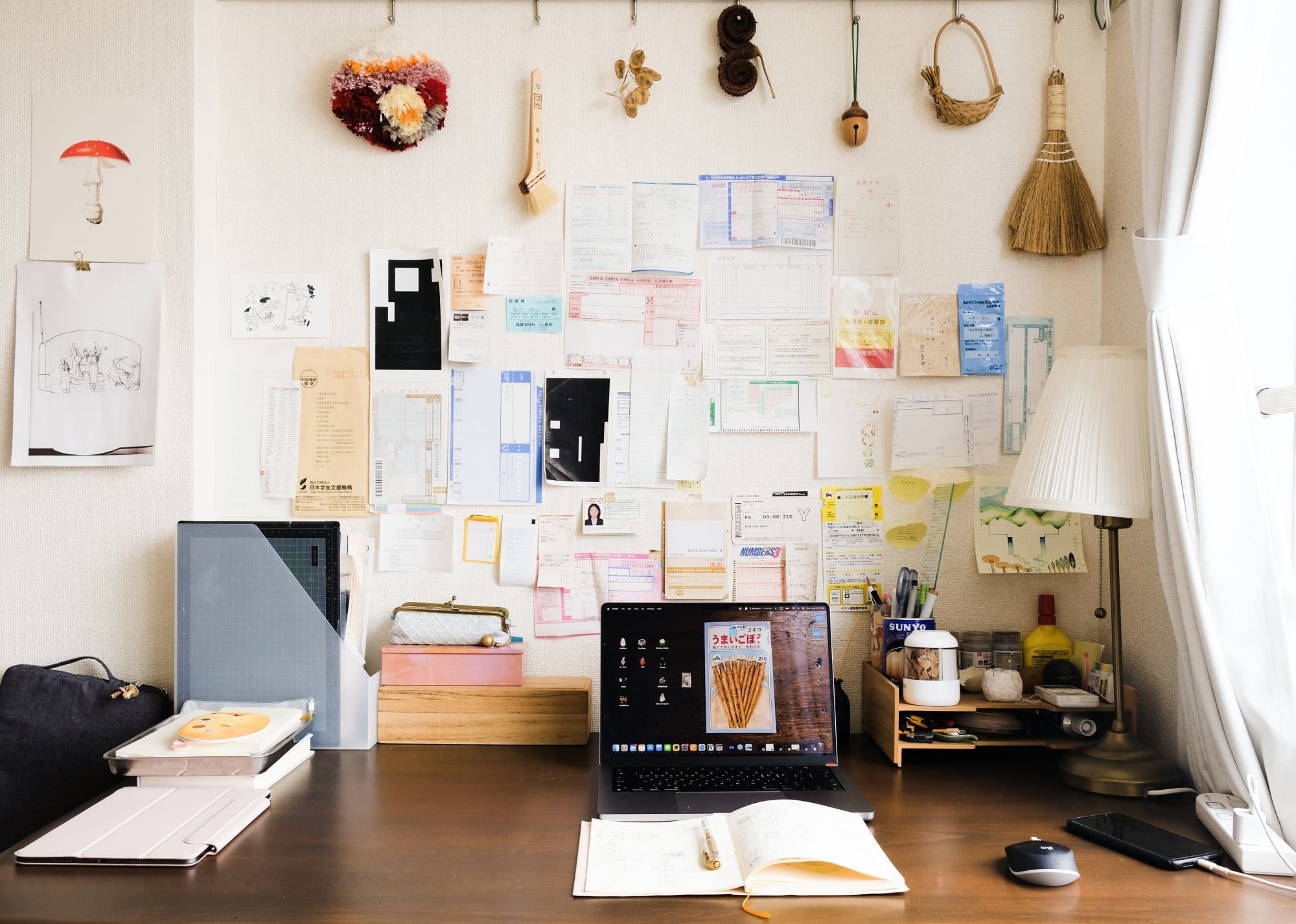 A neatly arranged desk setup with a laptop, open notebook, and stationery, set against a wall filled with documents and notes