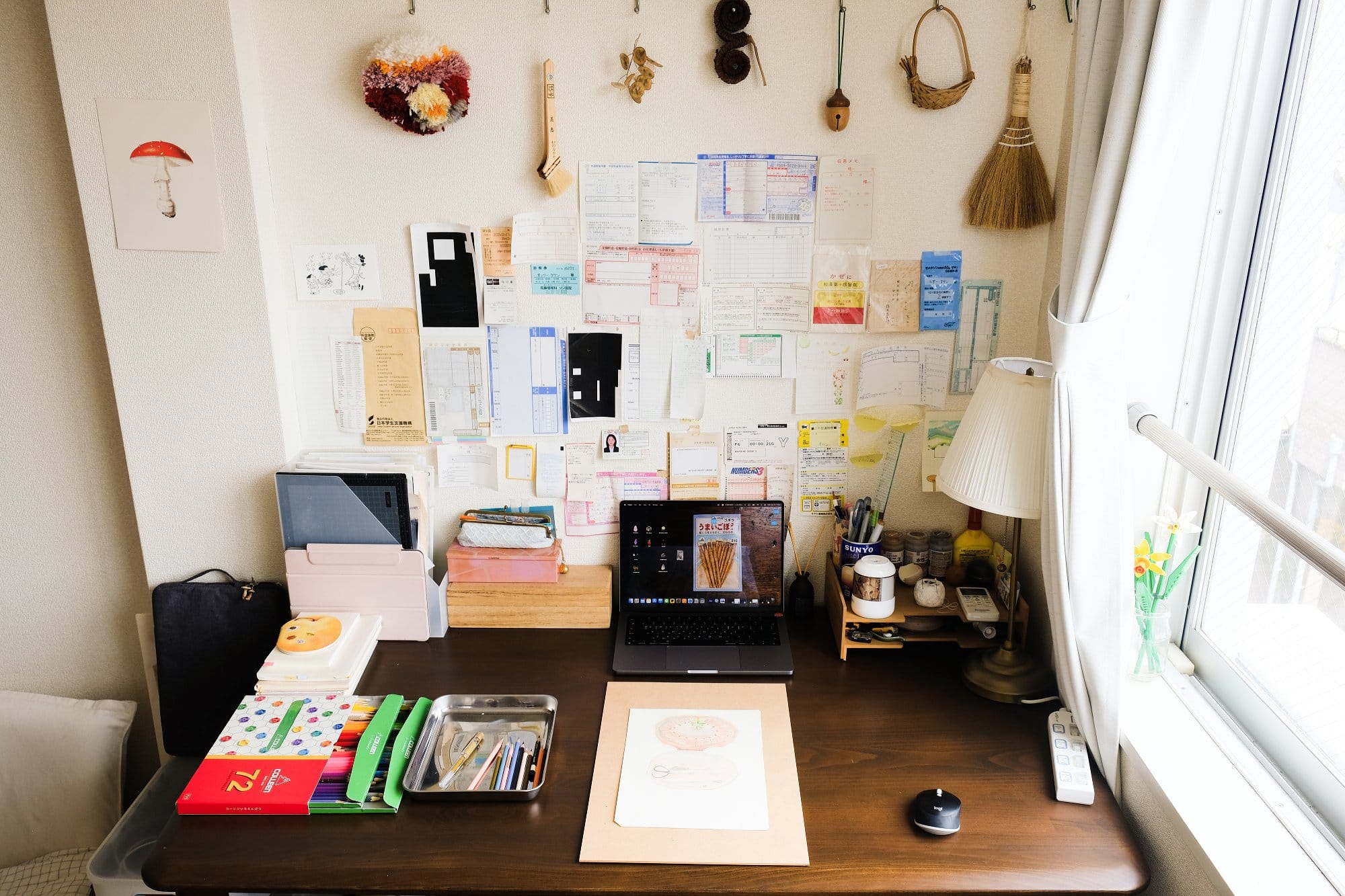 A tidy desk with a laptop, art supplies, and notebooks is set against a wall decorated with hand-tufted mini carpet, a traditional Japanese brush, dried plant items, a pine cone, an acorn-shaped folk toy, a basket, and a hand broom