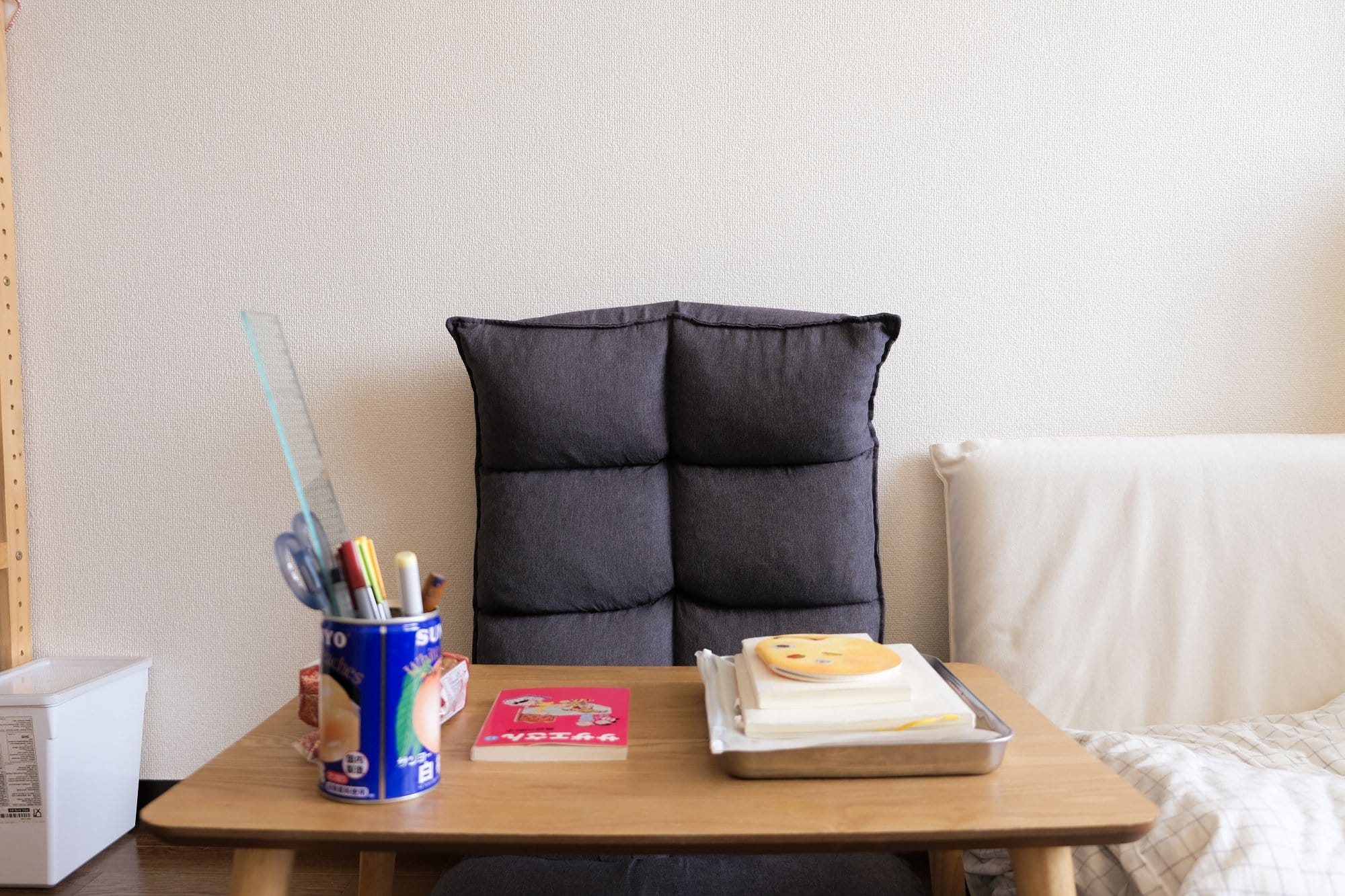 A small wooden table with a tin holding pens, a tray with books, and a cushion-backed chair positioned against a plain wall