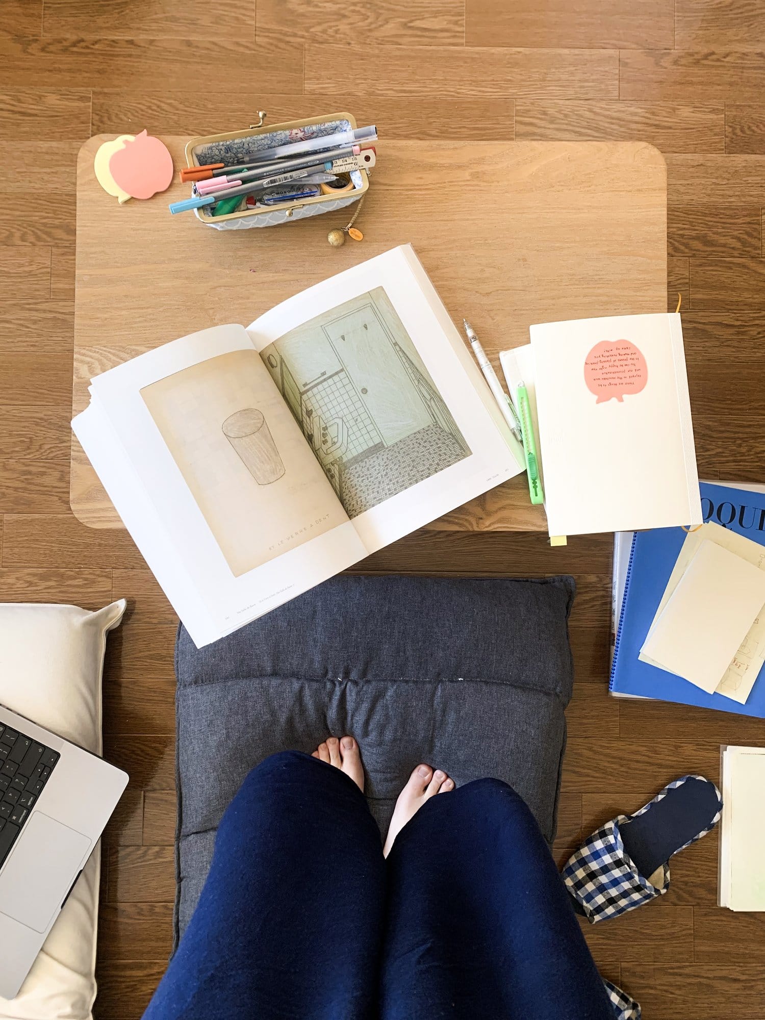 A top-down view of a person standing on a floor cushion, with bare feet visible, an open book, stationery, and a laptop placed on a low wooden table, surrounded by papers and slippers on the floor