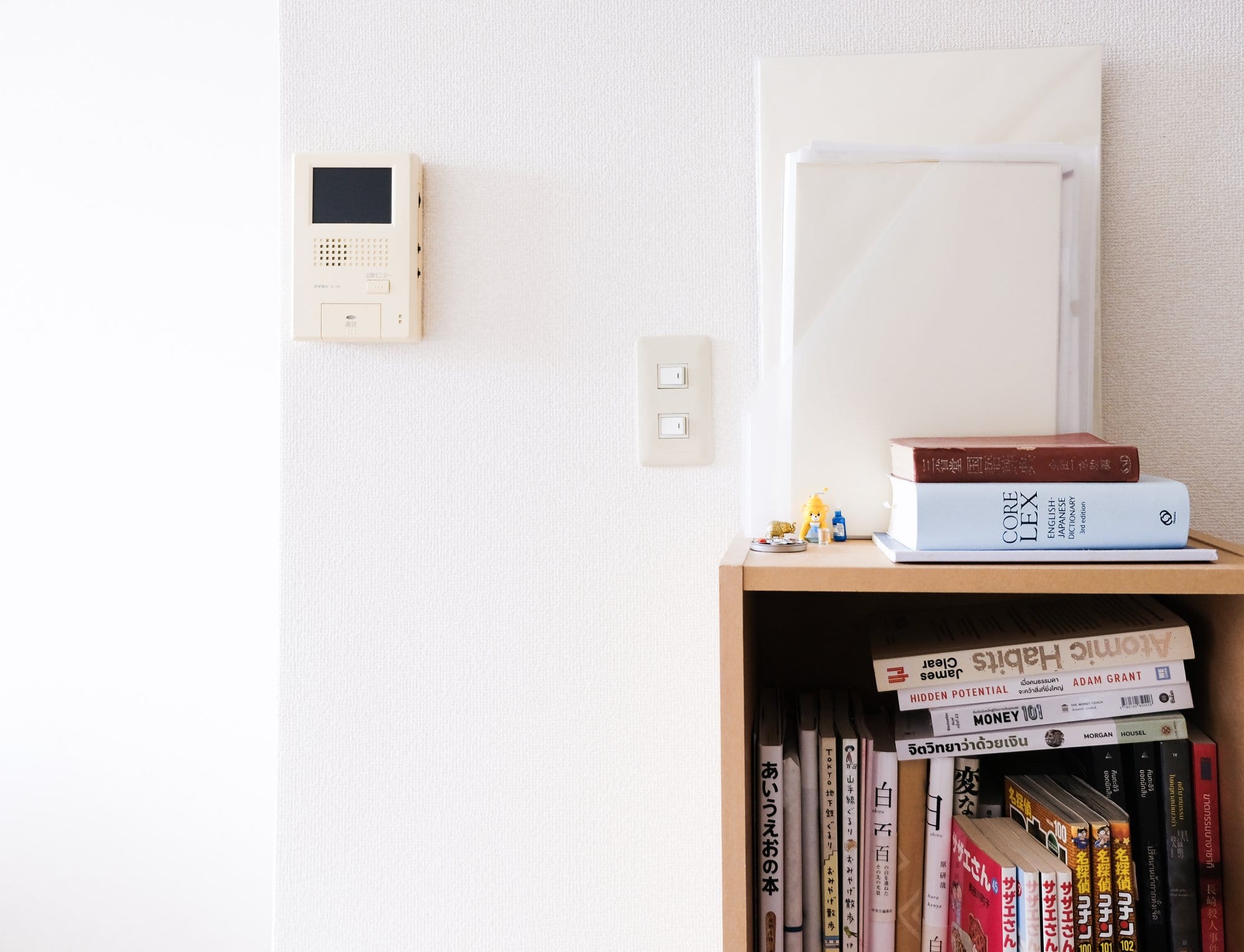 A bookshelf containing various books, including “Atomic Habits” and “Hidden Potential”, with a stack of folders and two figurines on top, next to a wall with a control panel and light switches