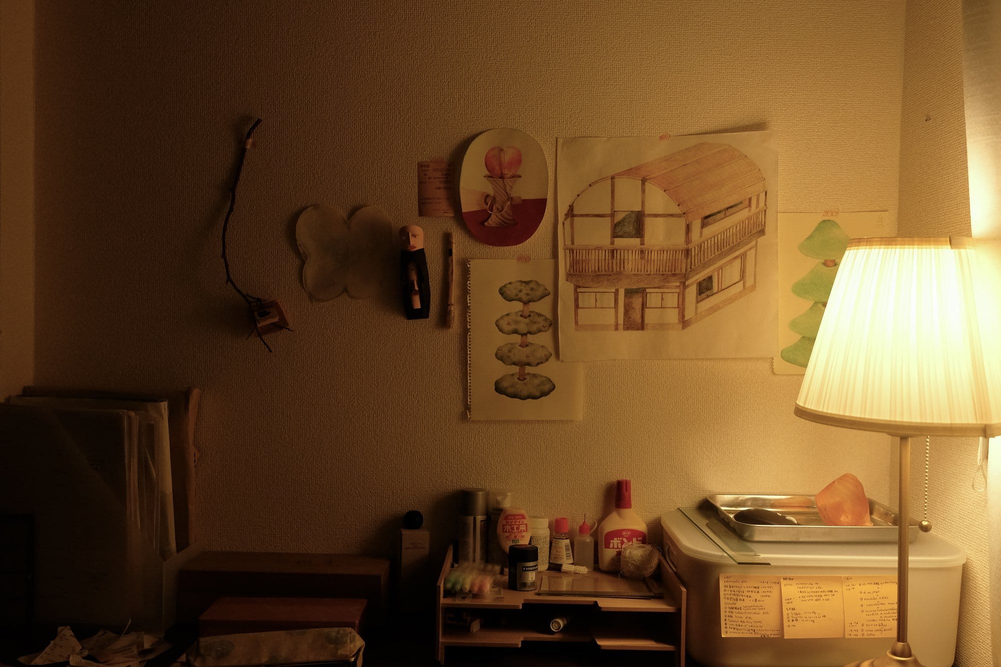 A cosy, softly lit corner with a desk, illuminated by a lamp, featuring art prints on the wall, small figurines, and neatly organised supplies and boxes