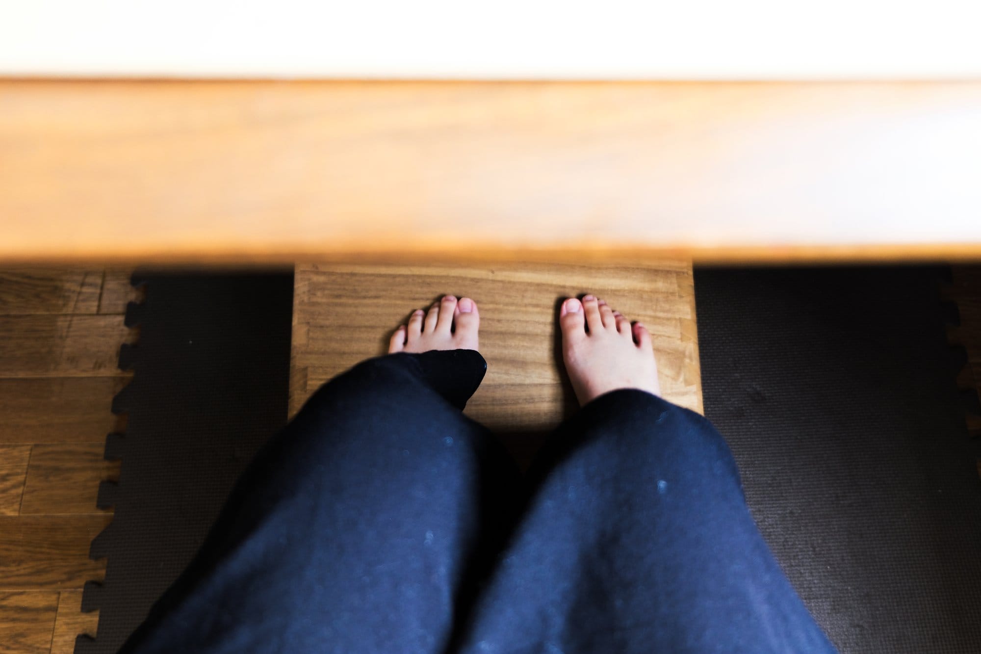 A view of bare feet resting on a wooden footrest, with dark trousers visible and the edge of a desk above