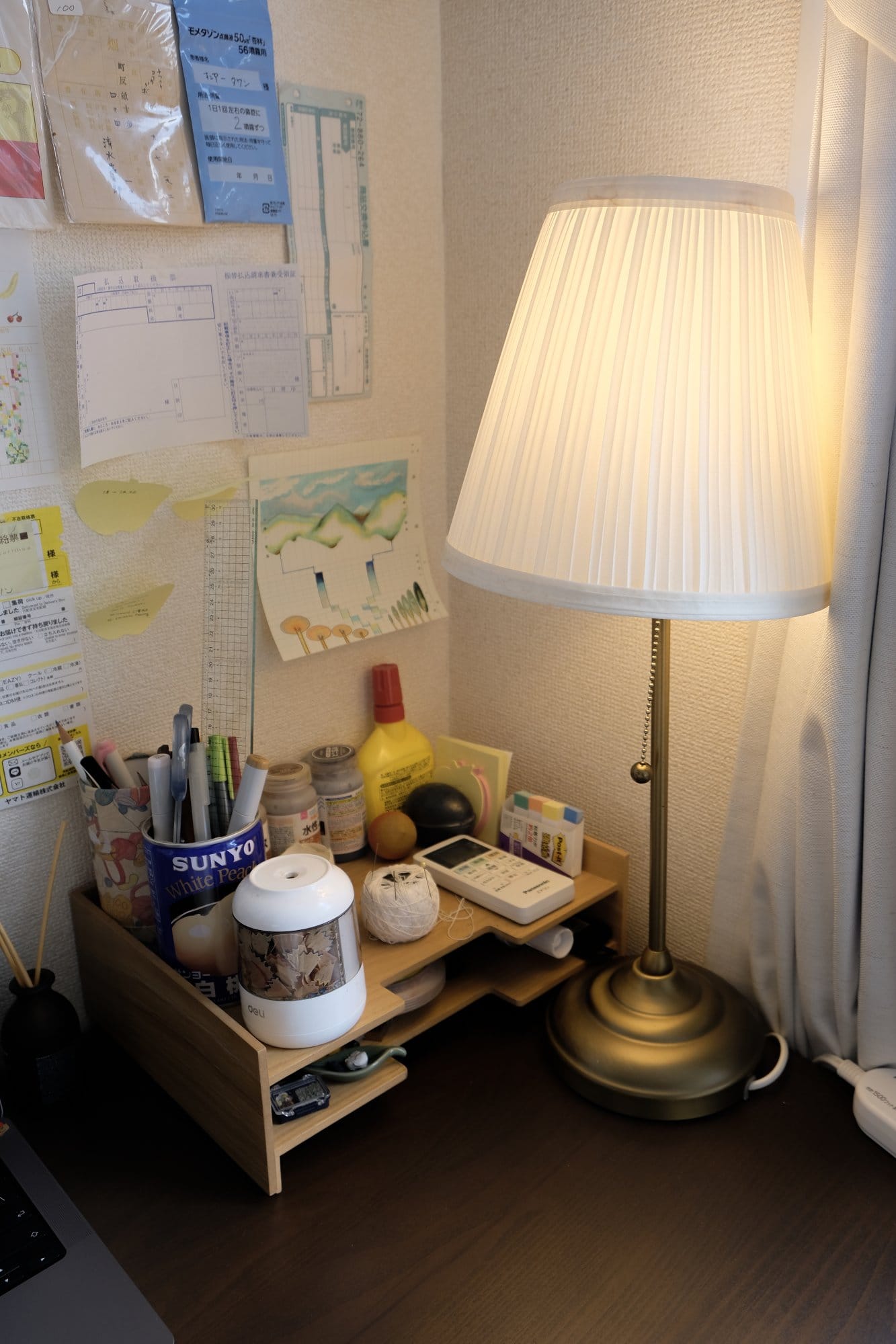 A desk corner with an IKEA ÅRSTID lamp, a wooden organiser filled with stationery and art supplies, and notes and drawings pinned to the wall above