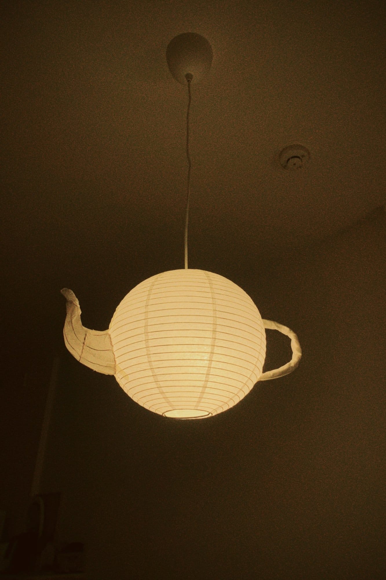 A ceiling lamp in the shape of a teapot, made from a paper lantern, softly glowing in a dimly lit room