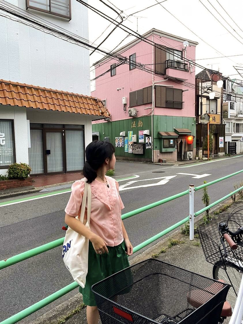 A photo of Thai-born, Japan-based artist Lily Ongartthaworn standing on a quiet street in Tokyo, wearing a pink blouse and green skirt, with colourful buildings in the background