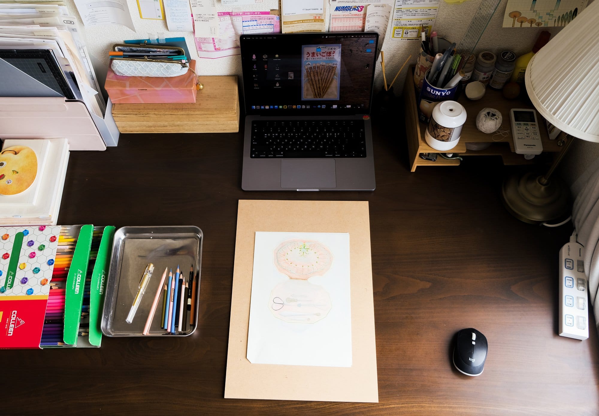 A workspace with a laptop, a Logitech mouse, pencils neatly placed in a tray, and an art project on a wooden board, surrounded by organised stationery and supplies on the desk