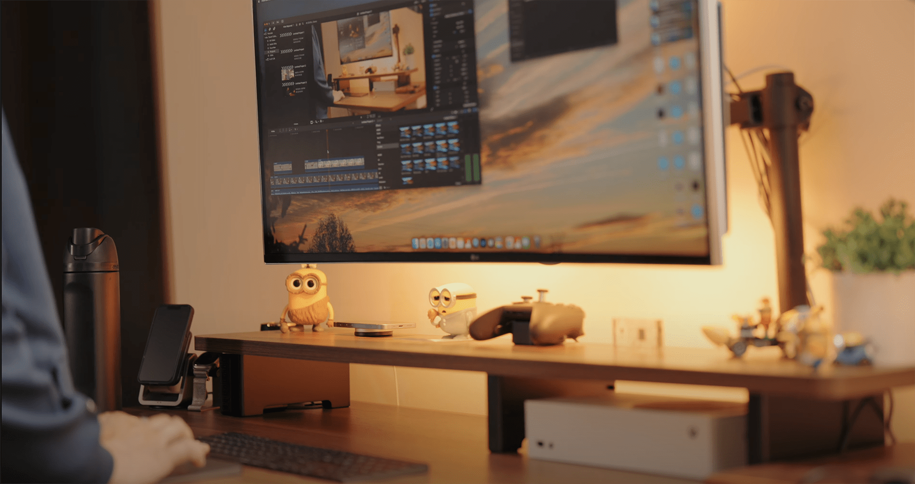 A home office workspace with an LG 38WN95C-W monitor displaying video editing software, an Xbox controller, and small decorations on a Vernal desk shelf, with a MacBook Pro and water bottle visible nearby