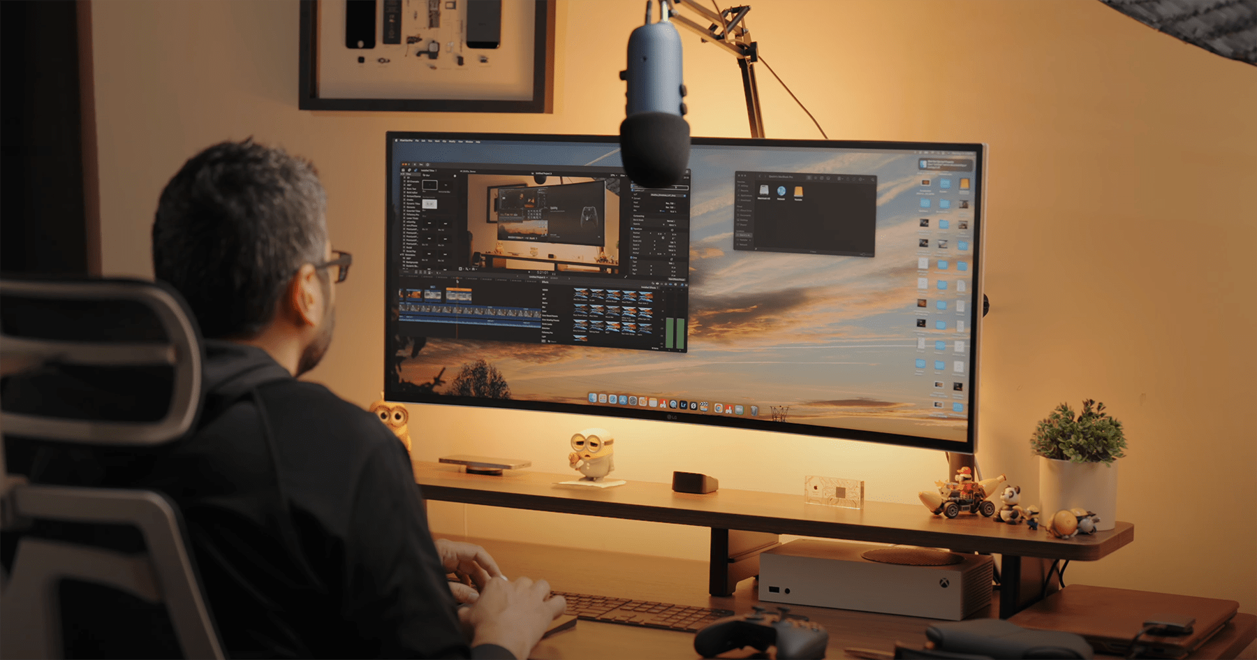 A person using a home office setup with an LG 38WN95C-W monitor on a Vivo Arm, a Blue Yeti microphone, a MacBook Pro, and an Xbox Series S on a Vernal desk shelf