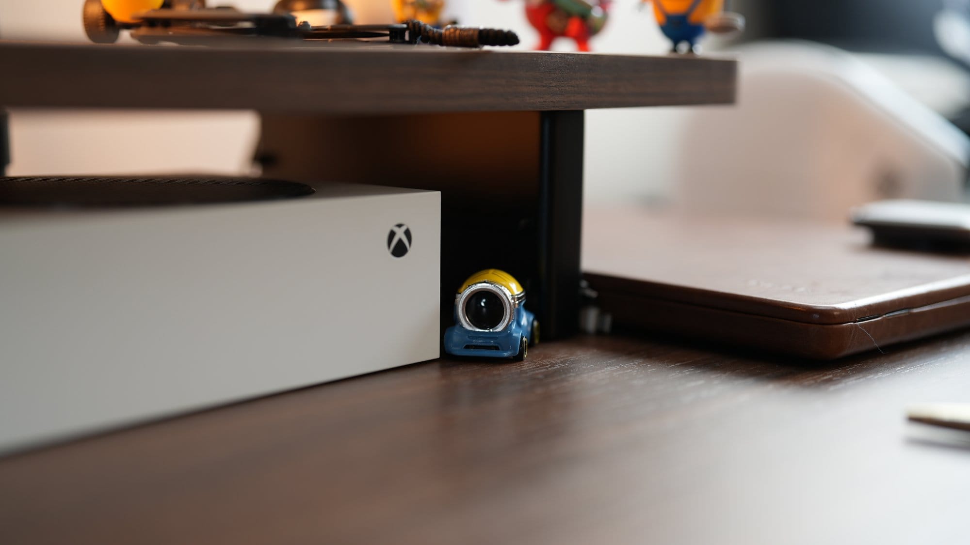 A close-up of an Xbox Series S console placed under a desk shelf, next to a small Minion toy figurine