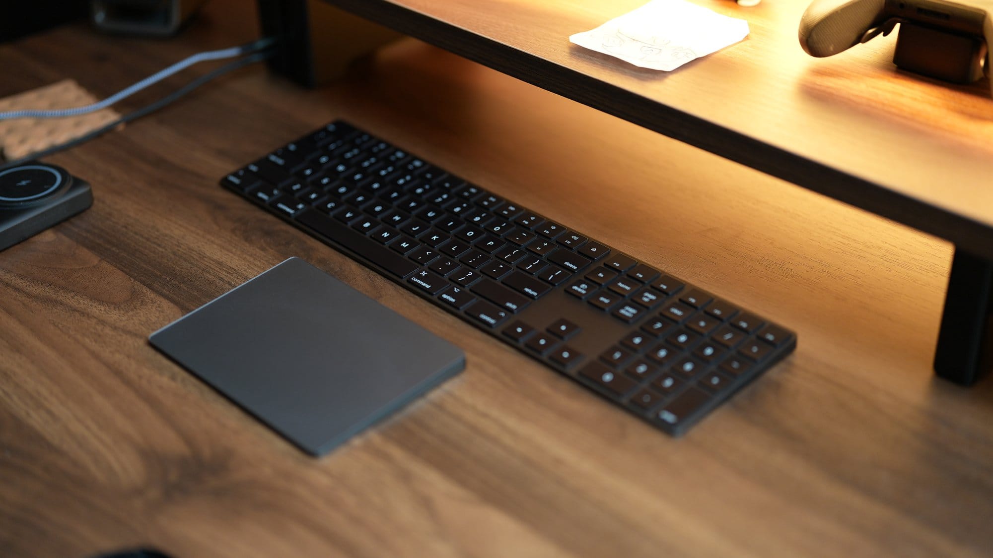 A close-up of an Apple Magic Keyboard and a Magic Trackpad placed on a wooden desk beneath a desk shelf